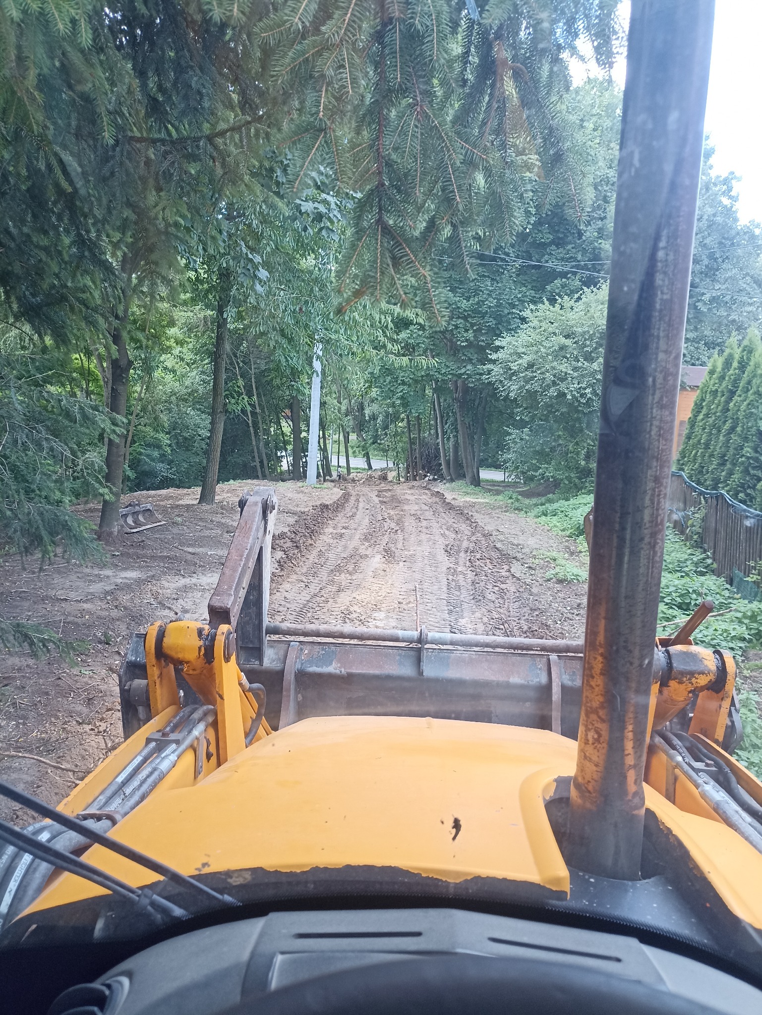 Widok z kabiny koparki na utwardzoną, błotnistą drogę leśną. Żółta maszyna gotowa do pracy, otoczona zielenią drzew.