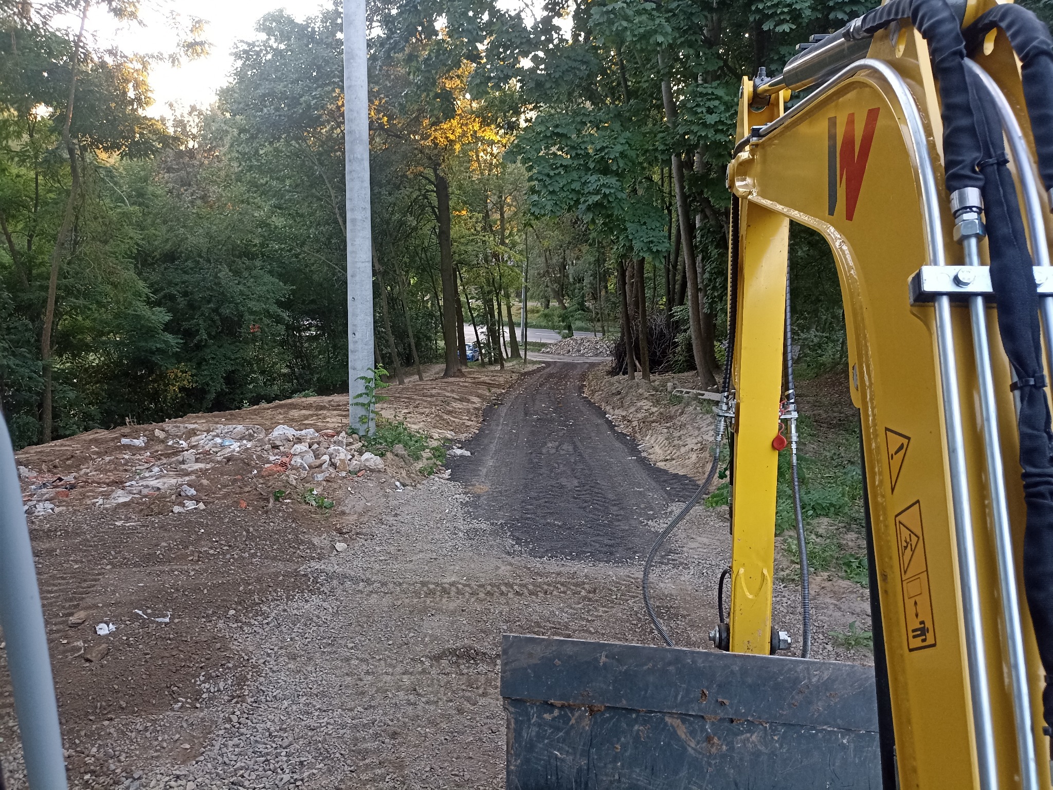 Żółta koparka na tle leśnej drogi w trakcie utwardzania. Widoczne ślady opon i świeżo rozsypany żwir. W tle drzewa i słup oświetleniowy.