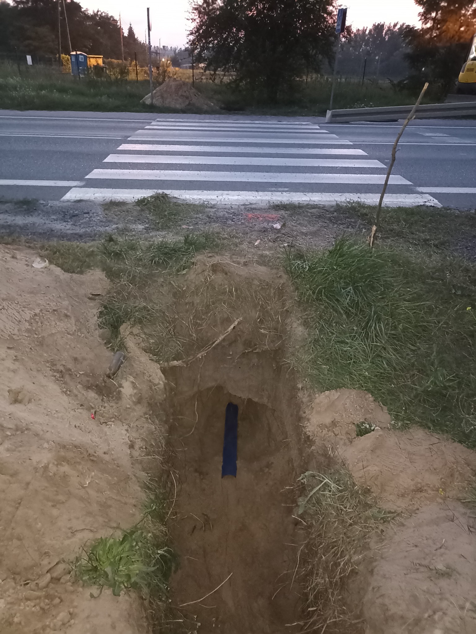 Wąski wykop w ziemi z widoczną niebieską rurą, położony obok drogi z przejściem dla pieszych. W tle widoczny przenośny toi-toi i hałda ziemi.