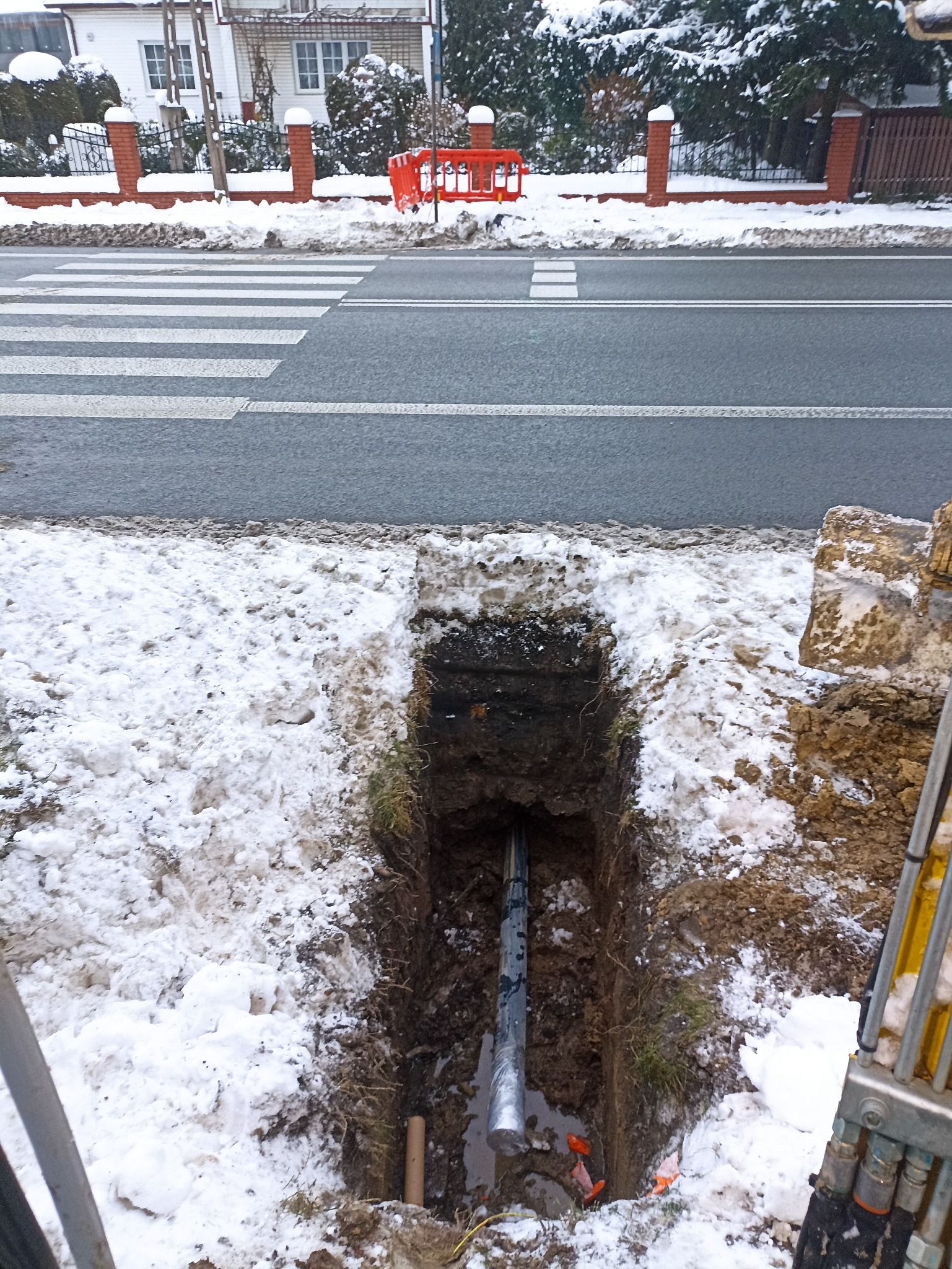 Zimowy wykop pod rurę przy drodze z przejściem dla pieszych. W wykopie widoczna rura, na ziemi śnieg. Na drugim planie zabudowania.