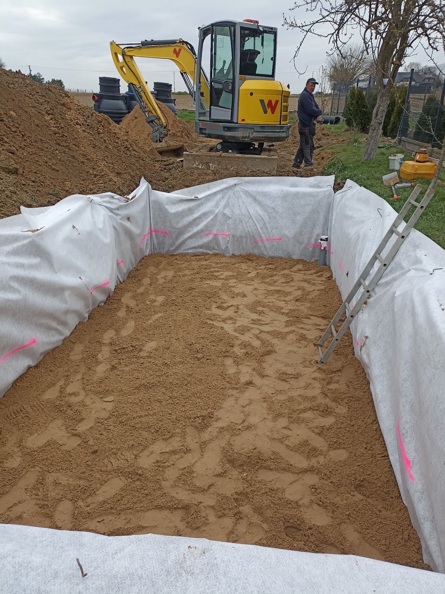 Wykop zabezpieczony geowłókniną, z piaskowym dnem. W tle minikoparka Wacker Neuson i czarne zbiorniki. Prace ziemne w toku, widoczna drabina.