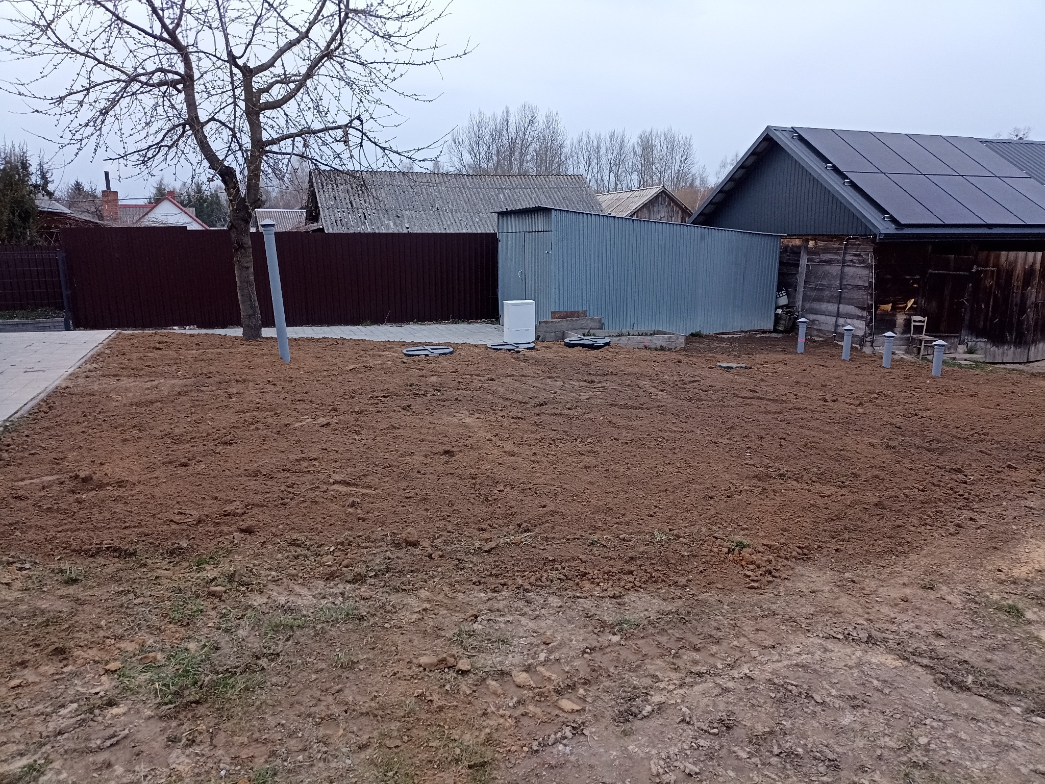 Przygotowany teren pod budowę z widocznymi rurami i studzienkami, w tle budynki gospodarcze i ogrodzenie. Ziemia wyrównana, gotowa do dalszych prac.