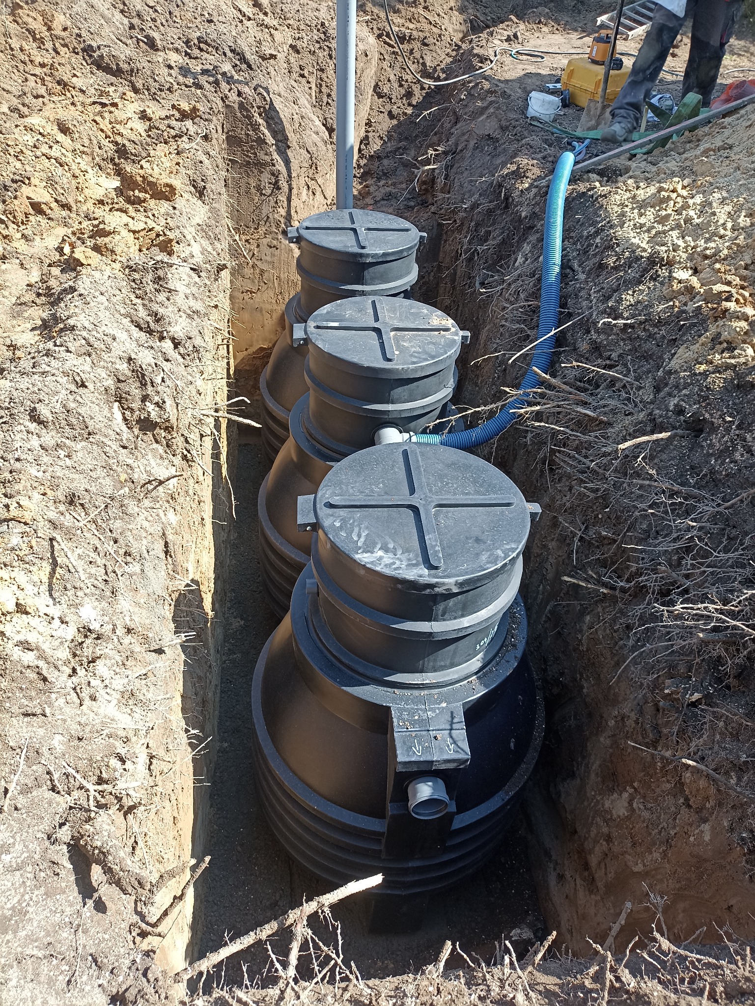 Wkopana w ziemię, czarna, trzykomorowa oczyszczalnia ścieków w Jabłonnie, z widocznymi rurami i wykopem. Widok z góry na instalację w ziemi.