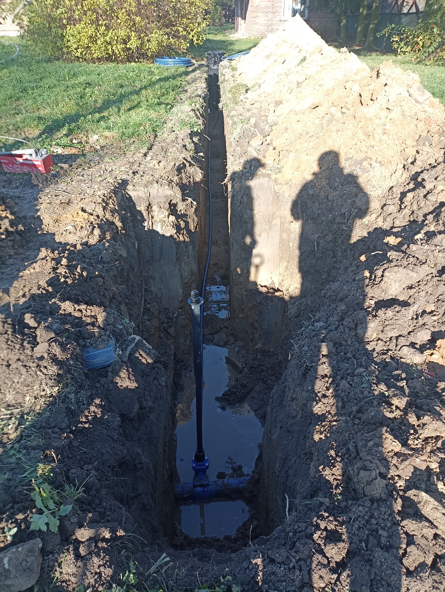 Wykopany rów z widoczną instalacją wodną, częściowo wypełniony wodą. Na brzegach ziemia i cień dwóch osób. W tle budynek i zieleń.