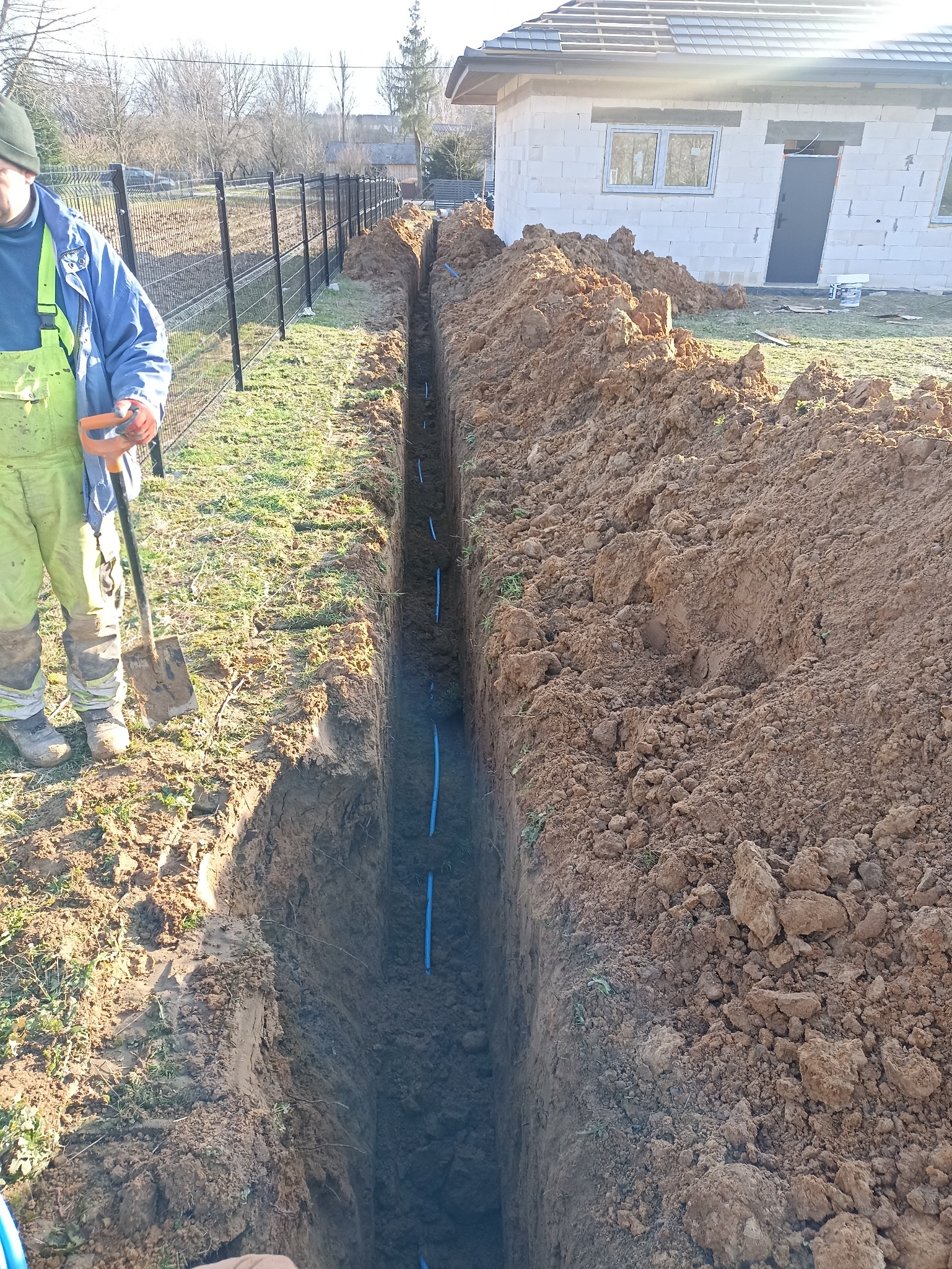 Wąski wykop z ułożonymi niebieskimi rurami, obok stoi pracownik z łopatą, w tle ogrodzenie i budynek w trakcie budowy. Widok z góry.