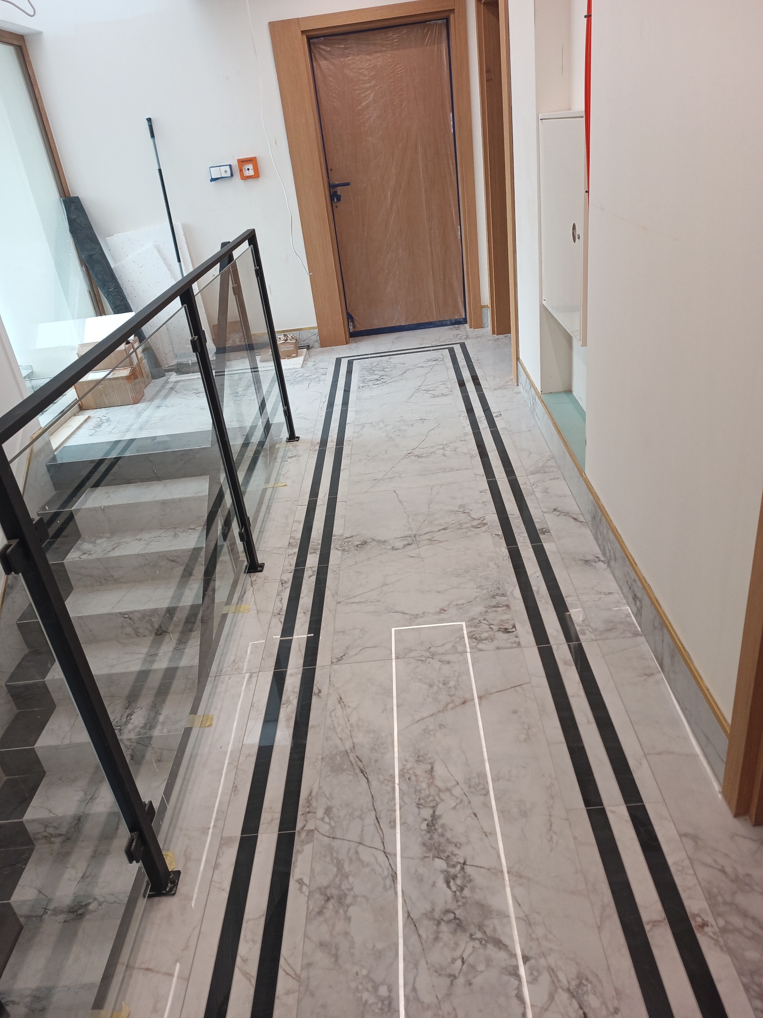 Marmurowe schody z czarnymi akcentami i szklaną balustradą w jasnym wnętrzu. Widoczne zabezpieczone drzwi i fragment okna. Wykończenie w stylu nowoczesnym.
