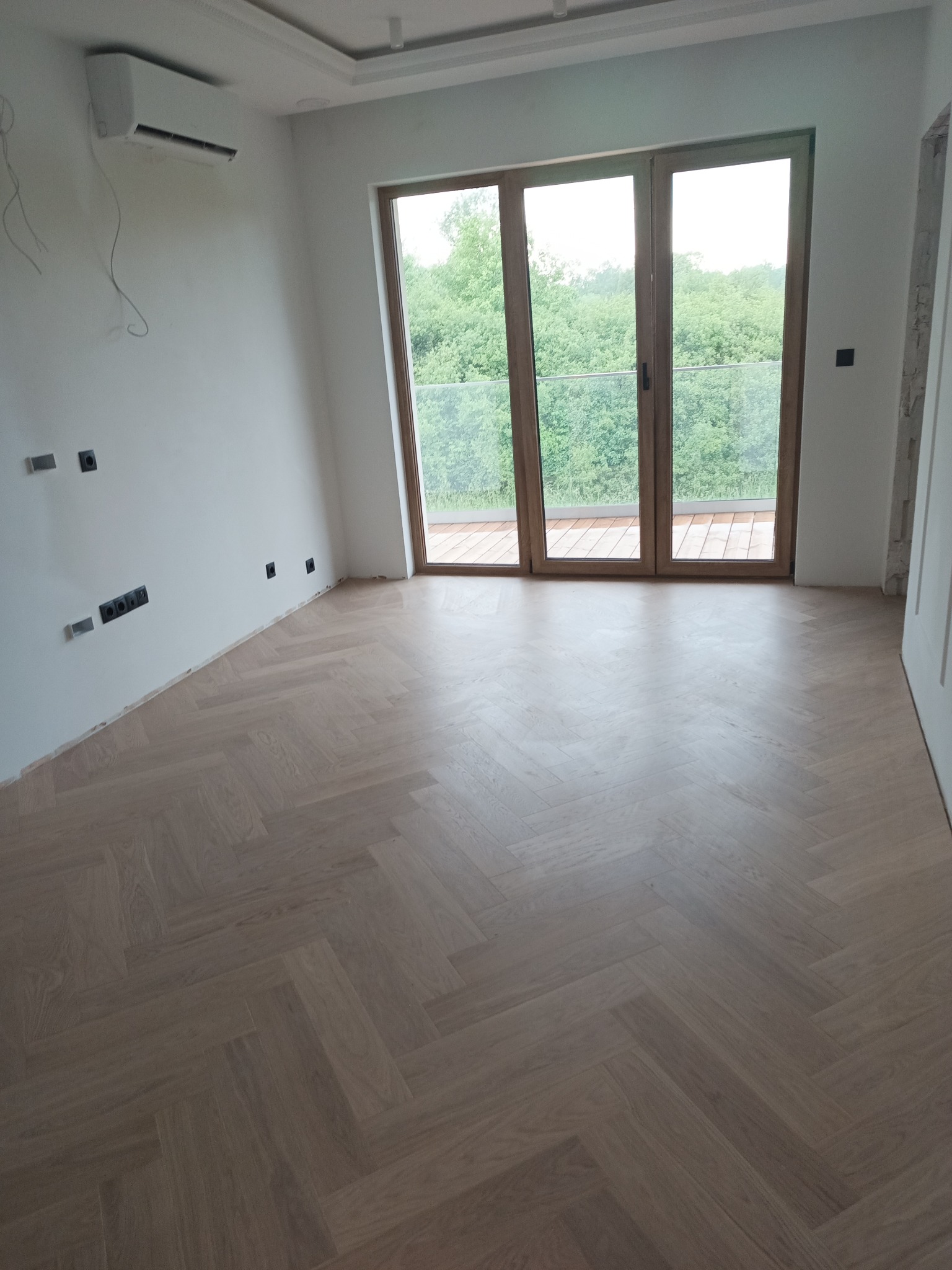 Jasny parkiet ułożony w jodełkę w nowym pokoju z dużym oknem balkonowym i widokiem na zieleń. Ściany białe, widoczne gniazdka. Klimatyzator na ścianie.