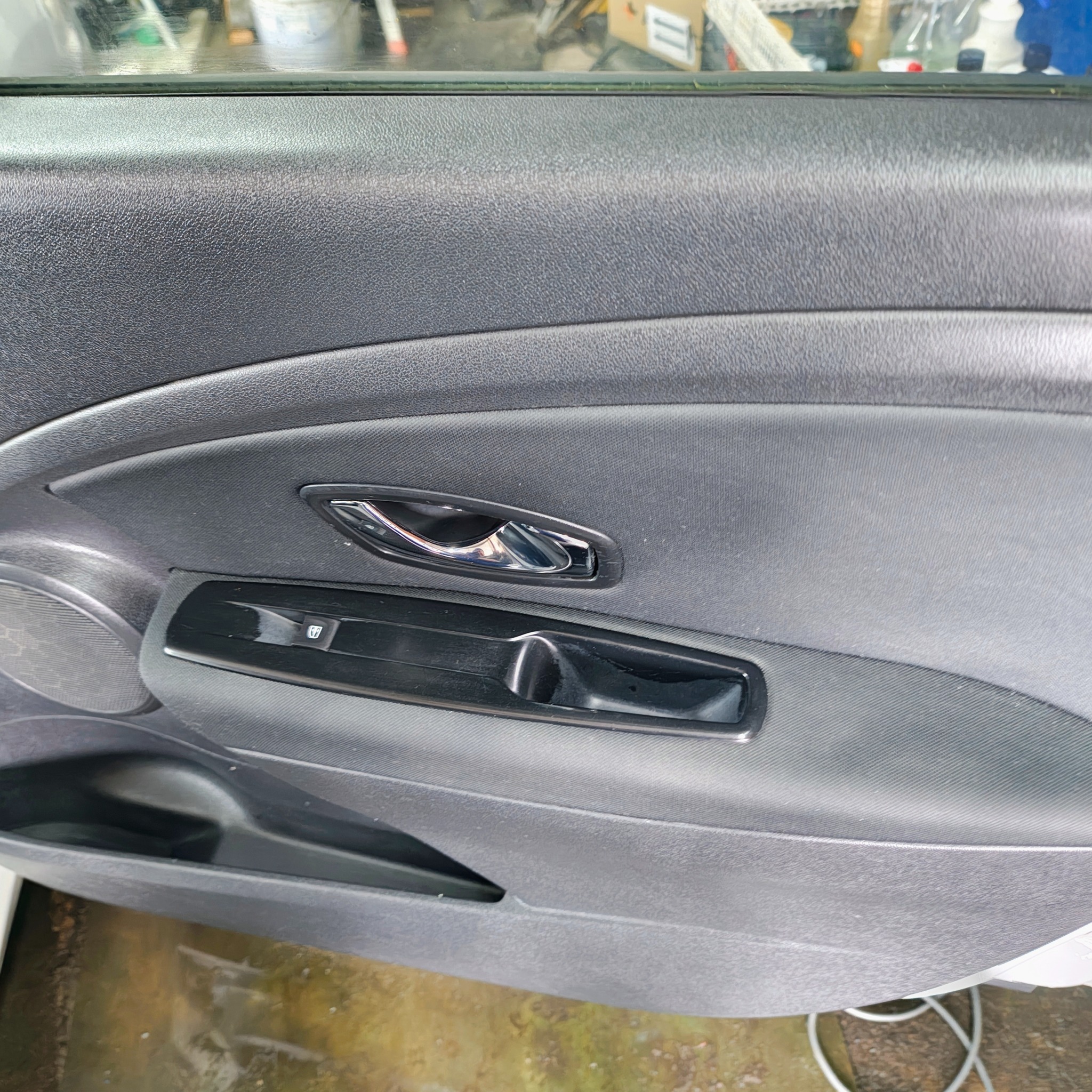 Szczegółowe ujęcie wewnętrznej strony drzwi samochodu, z widoczną chromowaną klamką, panelem sterowania szybami i głośnikiem. Teksturowana powierzchnia tworzywa.