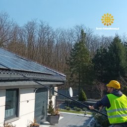 serwis-fotowoltaiczny.pl - Mężczyzna w kasku czyści panele słoneczne na dachu domu za pomocą długiego kija. Widoczny budynek, drzewa w tle i logo firmy serwis-fotowoltaiczny.pl.