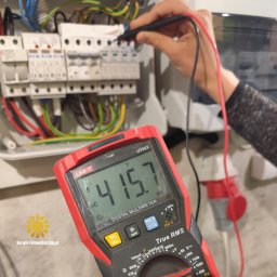 serwis-fotowoltaiczny.pl - Pomiar napięcia 415.7V miernikiem Uni-T UT89X na instalacji elektrycznej z bezpiecznikami. Widoczne przewody i gniazdo. Logo Serwis-Fotowoltaiczny.pl w lewym dolnym rogu.