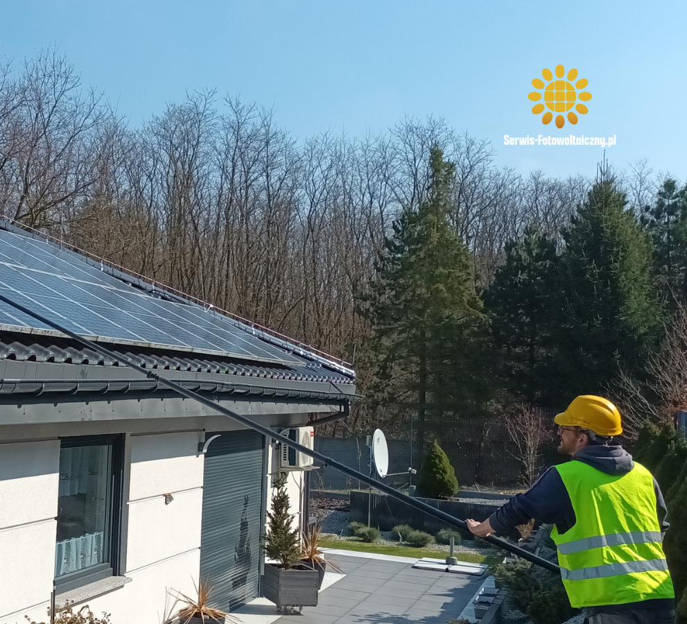 Mężczyzna w kasku czyści panele słoneczne na dachu domu za pomocą długiego kija. Widoczny budynek, drzewa w tle i logo firmy serwis-fotowoltaiczny.pl.