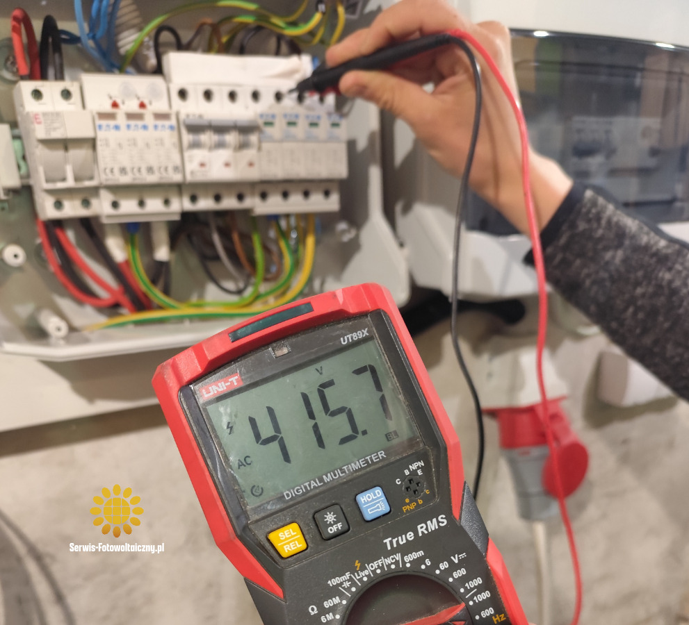 Pomiar napięcia 415.7V miernikiem Uni-T UT89X na instalacji elektrycznej z bezpiecznikami. Widoczne przewody i gniazdo. Logo Serwis-Fotowoltaiczny.pl w lewym dolnym rogu.