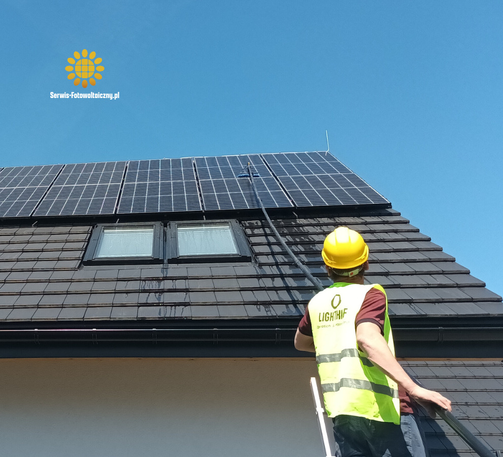 Czyszczenie paneli słonecznych na dachu domu przez pracownika w kasku i kamizelce odblaskowej przy użyciu długiej szczotki teleskopowej. Błękitne niebo w tle.