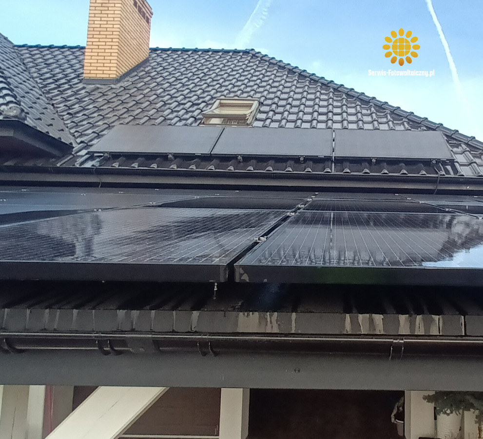 Czarne panele fotowoltaiczne zamontowane na ciemnym, spadzistym dachu domu z ceglanym kominem i oknem dachowym w słoneczny dzień. Widoczny fragment rynny.