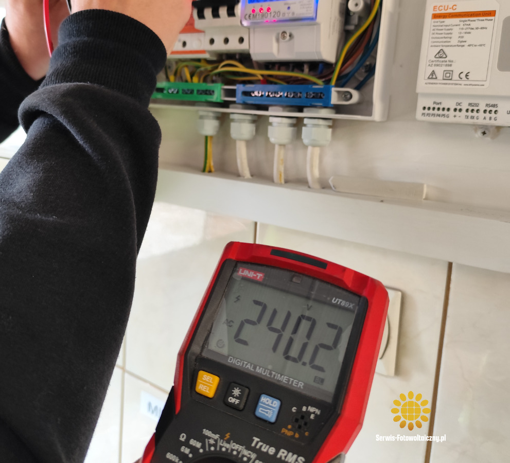 Elektryk mierzy napięcie 240.2V multimetrem UNI-T UT89X w skrzynce z bezpiecznikami i przewodami. Widoczny licznik CEM190120.