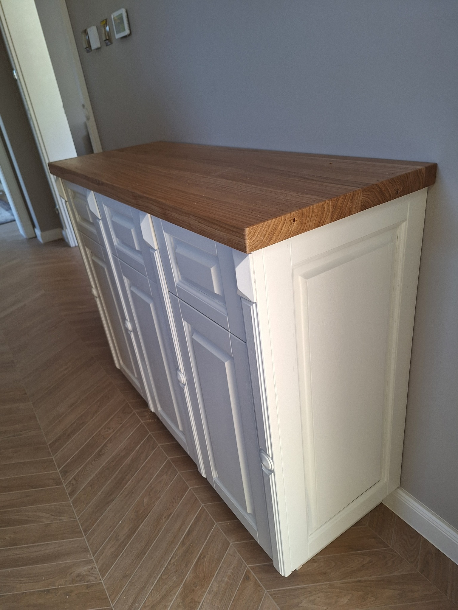 Biała szafka z drewnianym blatem, widoczna z boku na tle szarej ściany i podłogi w jodełkę. Klasyczny styl mebla z frezowanymi frontami.