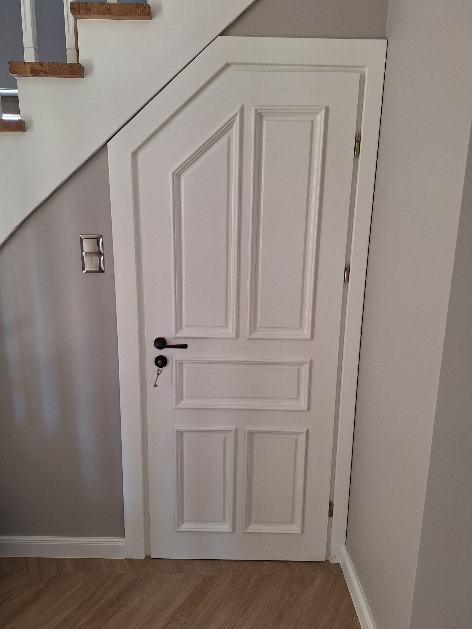 Białe, frezowane drzwi pod schodami, z czarną klamką i kluczem w zamku. Widoczny fragment schodów i podłoga z paneli. Szara ściana z włącznikiem światła.