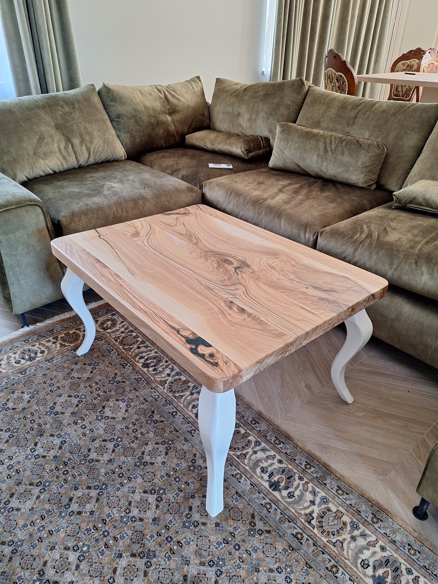 Stolik kawowy z blatem z jasnego drewna o unikalnym wzorze słojów i białymi, giętymi nogami. Na tle dywanu i narożnika w odcieniach zieleni.