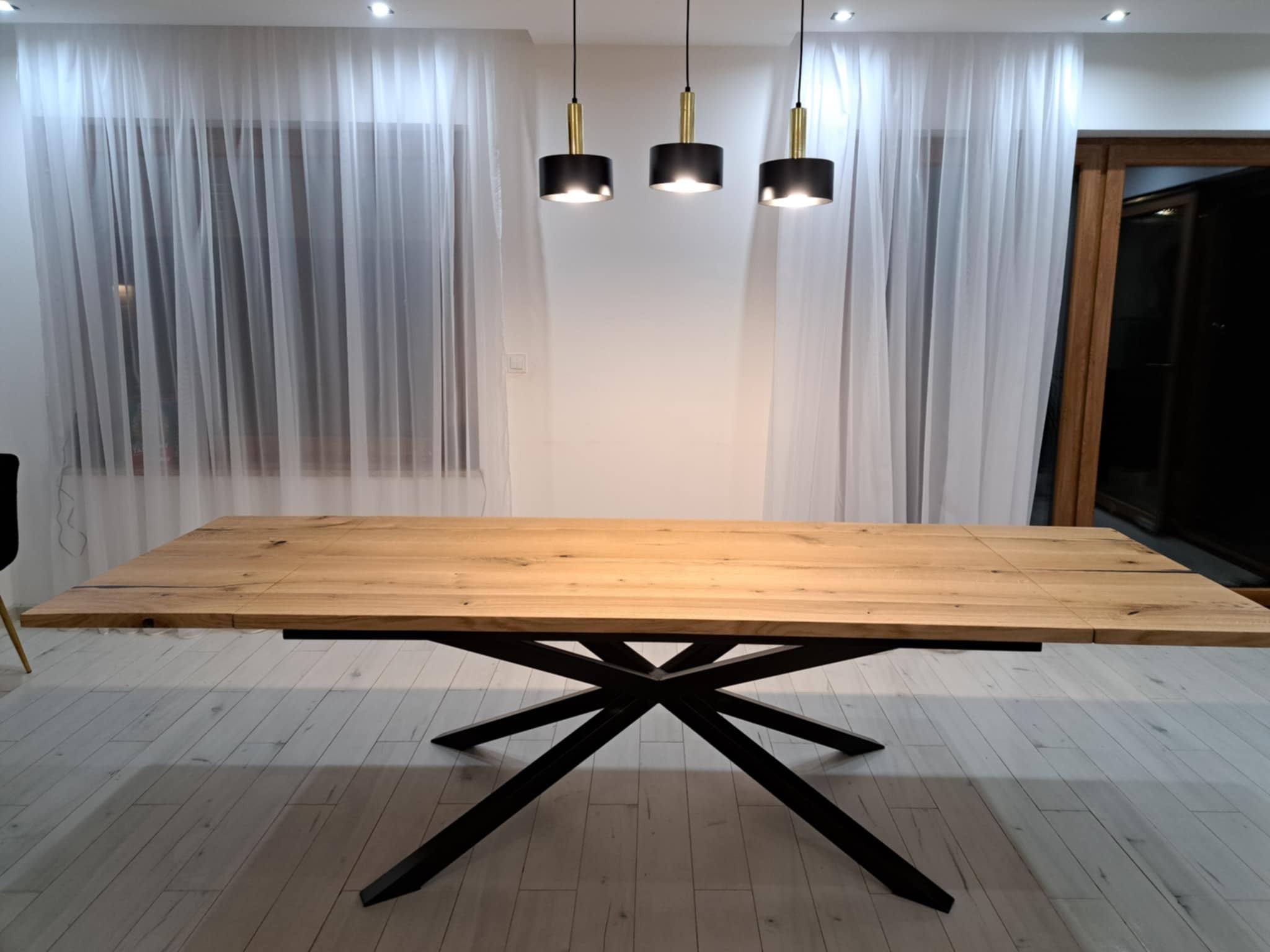 Rozkładany stół z blatem z jasnego drewna i czarną, geometryczną podstawą, oświetlony trzema wiszącymi lampami w nowoczesnym wnętrzu.