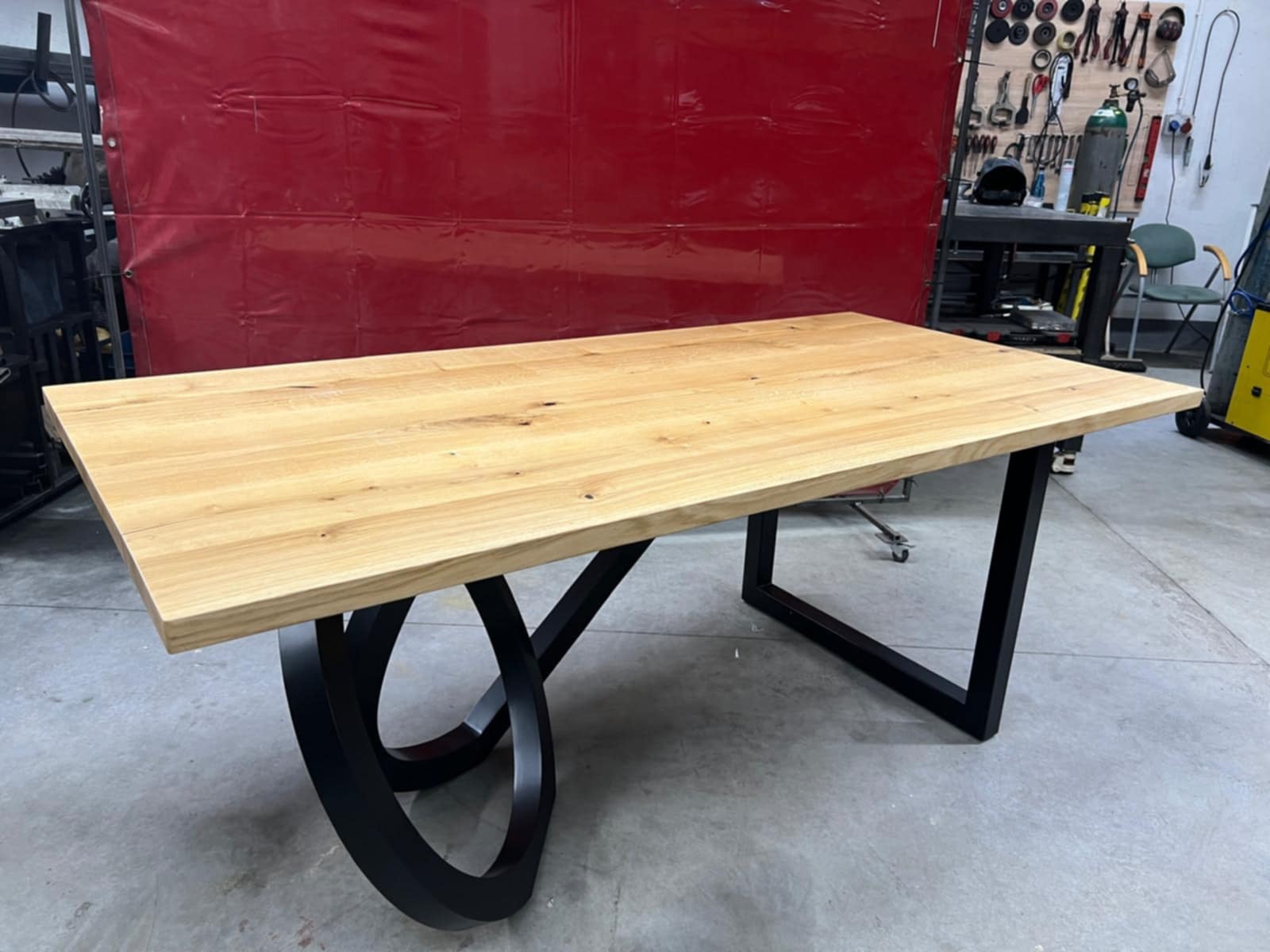 Stół z jasnego drewna z czarnymi, metalowymi nogami o unikalnym kształcie, ustawiony w warsztacie z czerwonym tłem i narzędziami.