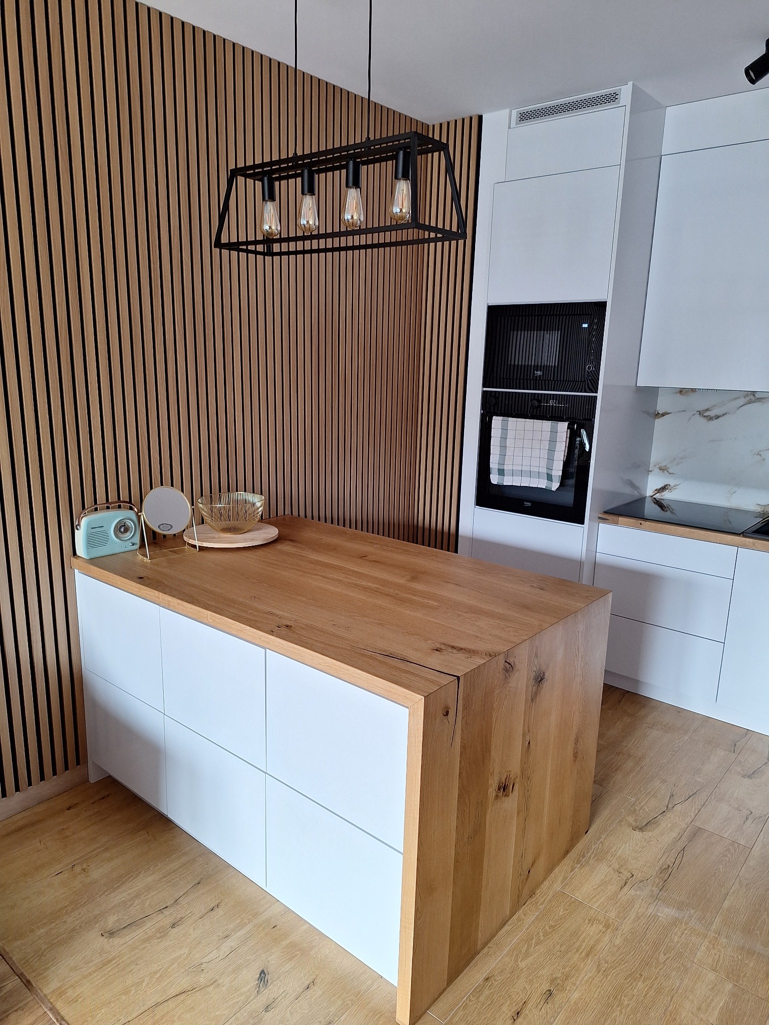 Nowoczesna kuchnia z drewnianą wyspą i lamelami na ścianie. Minimalistyczny design, białe szafki, czarne oświetlenie i elementy dekoracyjne.