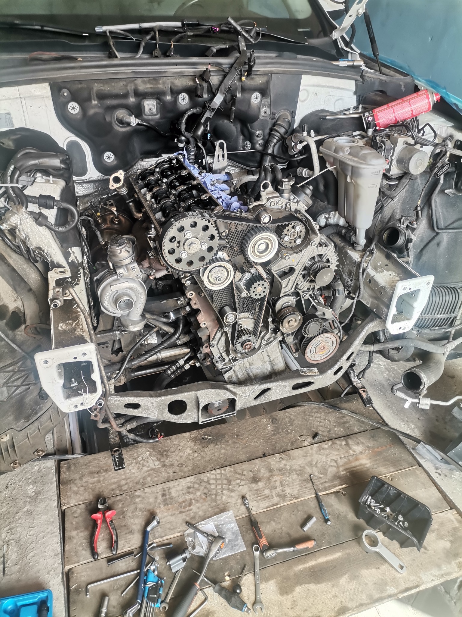 Widok z góry na rozmontowany silnik samochodowy z widocznymi narzędziami na drewnianym stole warsztatowym, ukazujący etap prac naprawczych.