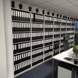 Taxxa Przyjazna Księgowa Biuro Rachunkowe Gdynia - Biuro rachunkowe: regał z segregatorami oznaczonymi numerami, rośliny doniczkowe na górze, biurka z komputerami w tle. Porządek w dokumentacji księgowej.