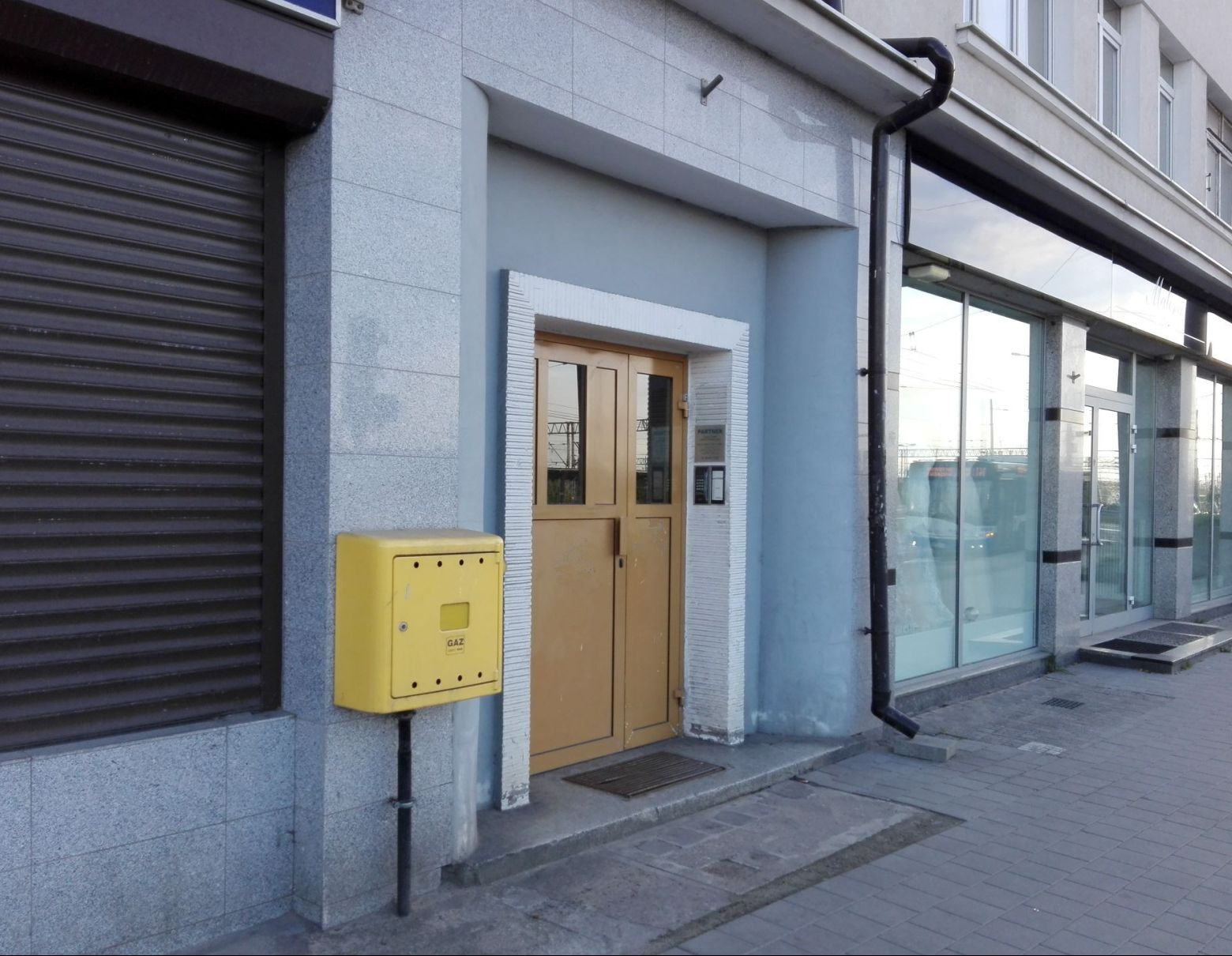 Wejście do biura z żółtą skrzynką gazową po lewej, szare płytki na elewacji i ciemne rolety obok. W tle widoczne okna z odbiciami ulicy.