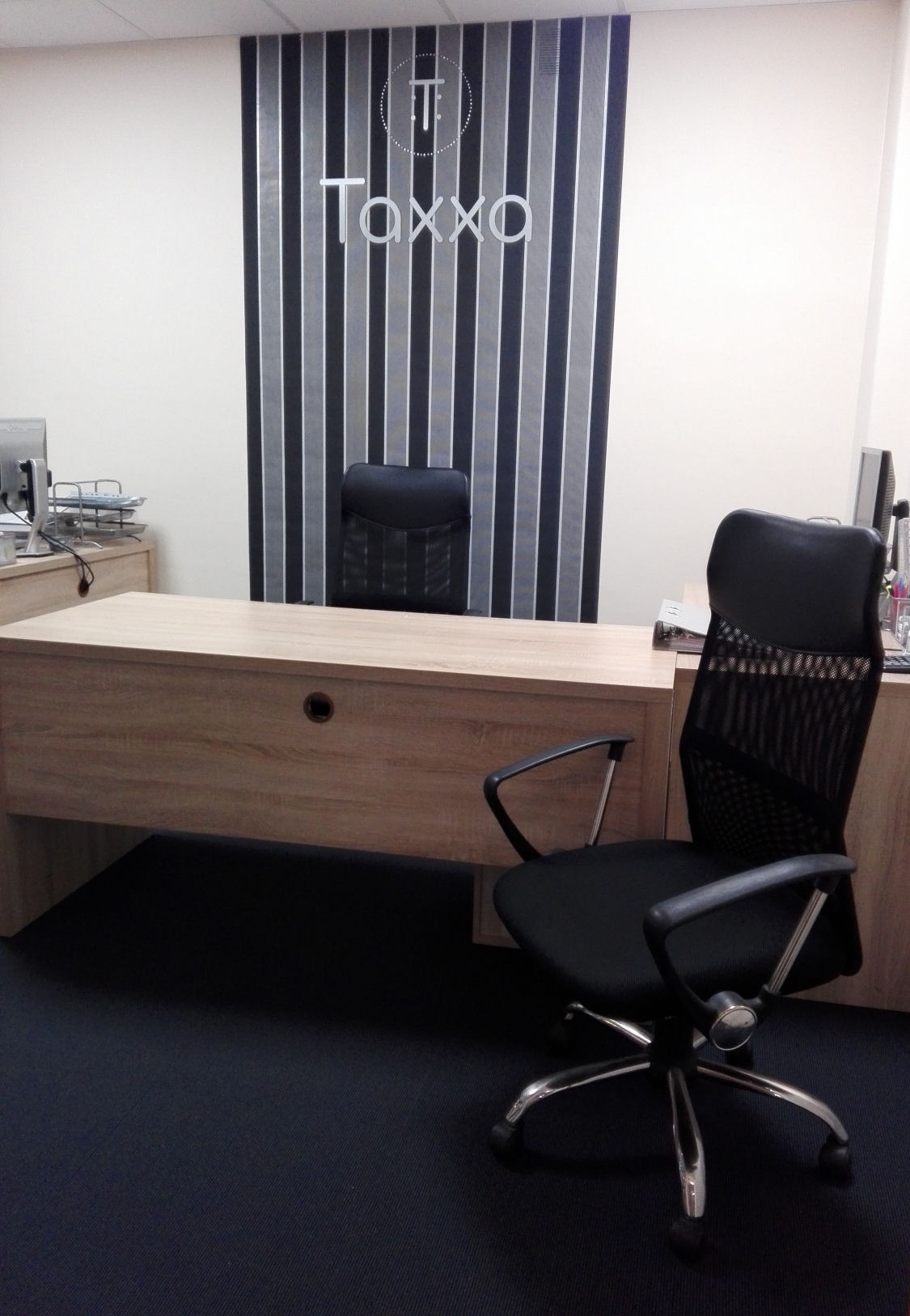 Nowoczesne biuro rachunkowe Taxxa: biurko z krzesłem na tle ściany z logo firmy, stonowana kolorystyka, minimalizm, czarne krzesło biurowe.