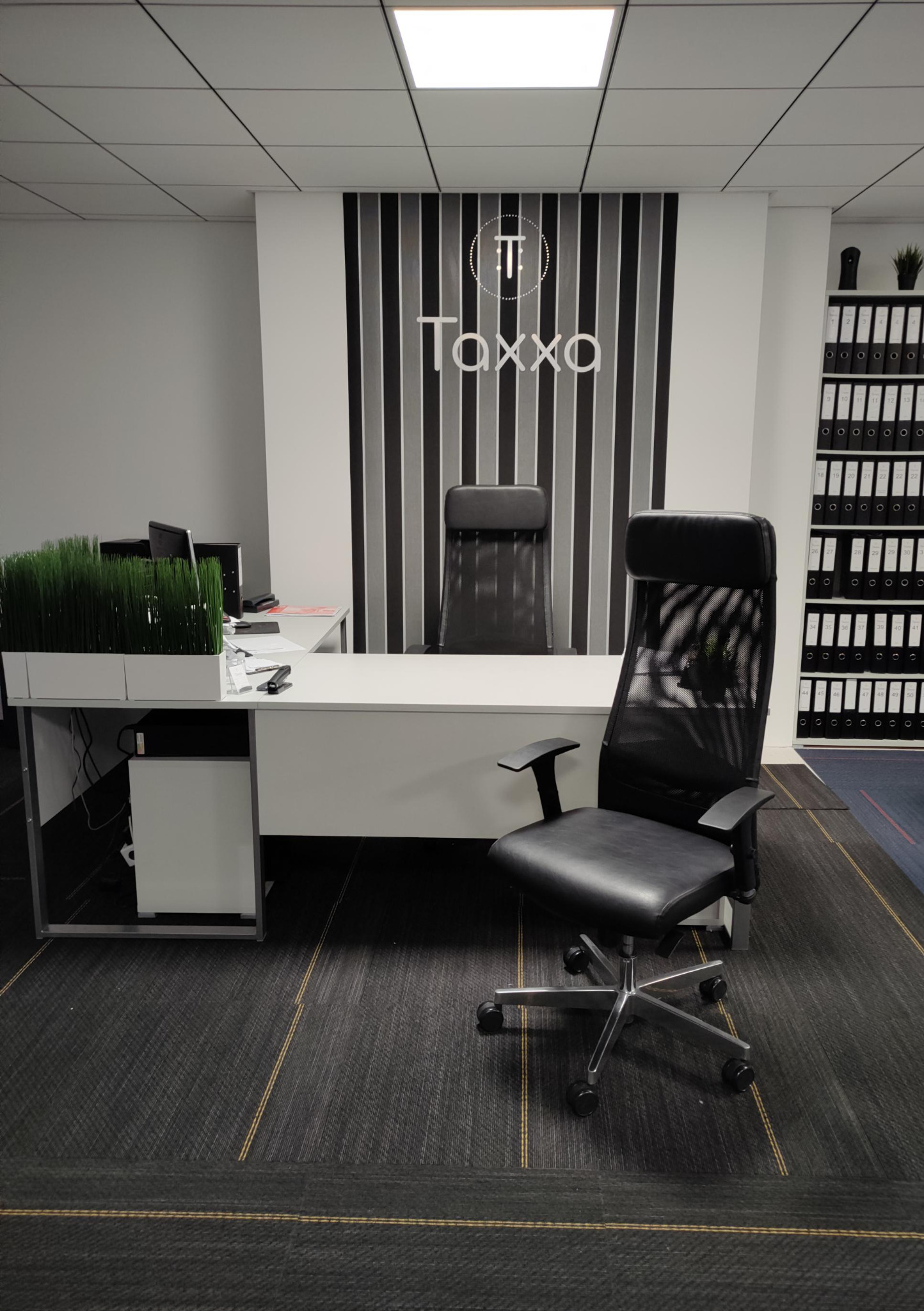 Nowoczesne biuro rachunkowe: minimalistyczny wystrój z biurkiem, krzesłem obrotowym i regałem na dokumenty. Logo firmy Taxxa na ścianie.