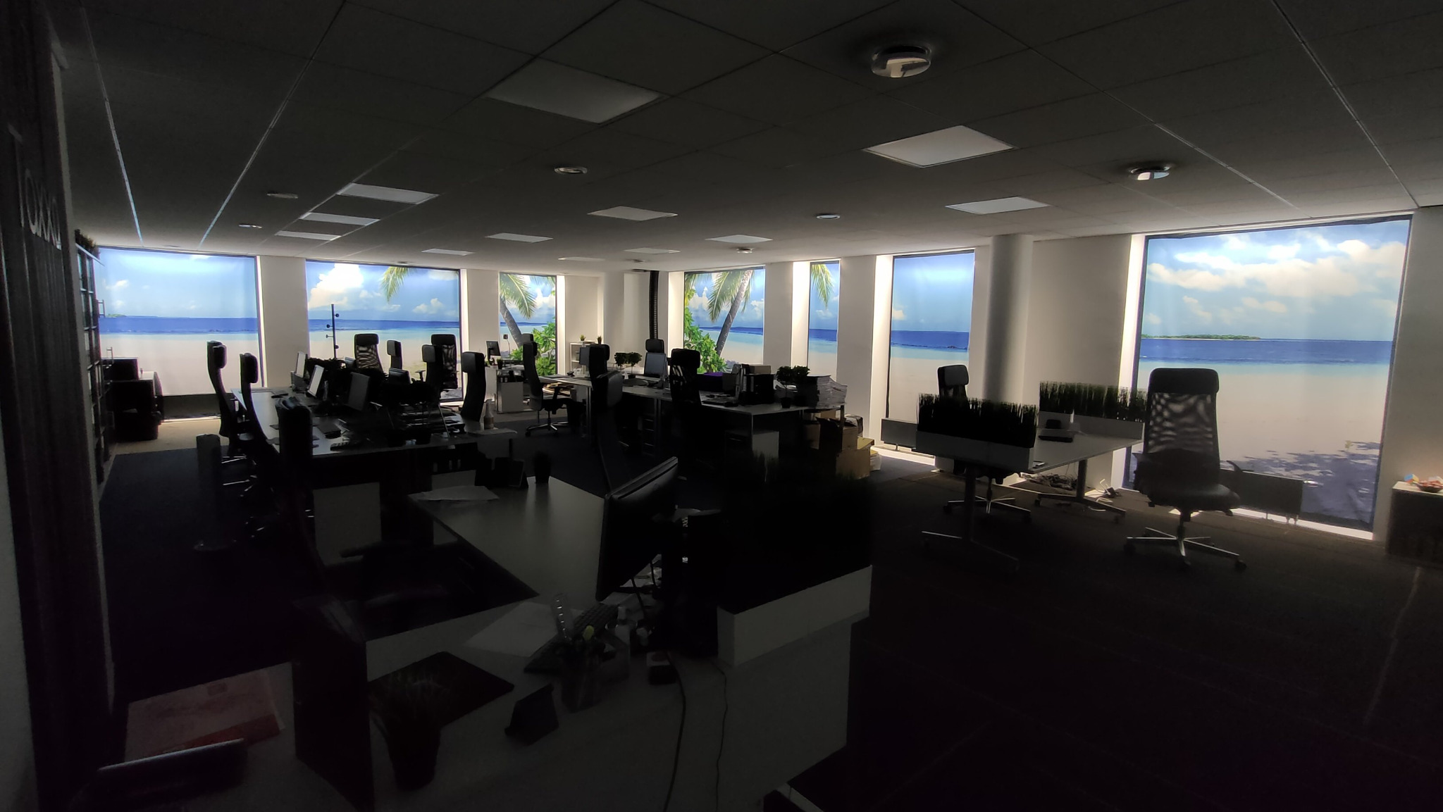 Nowoczesne biuro z widokiem na plażę. Przestronne wnętrze z biurkami i krzesłami, duże okna z fototapetami przedstawiające tropikalny krajobraz morski.