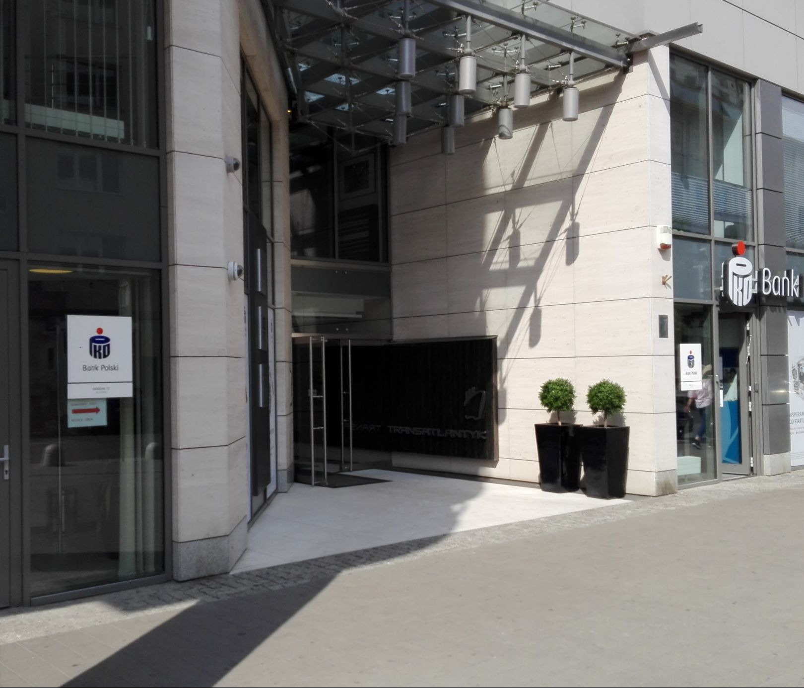 Wejście do biurowca z logo banku PKO BP i nazwą Transatlantyk. Dwie donice z roślinami przed wejściem. Szklane zadaszenie nad drzwiami.