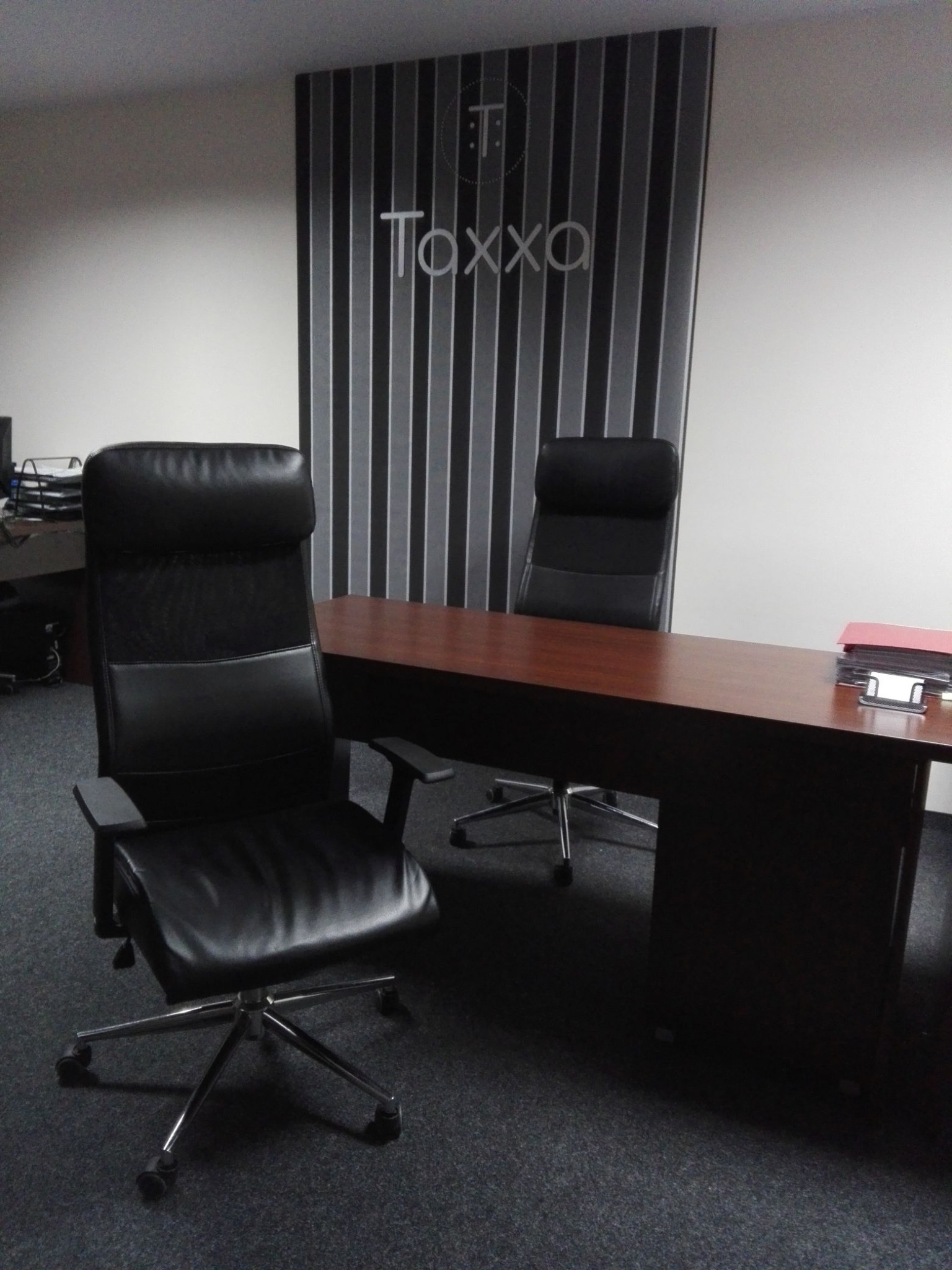 Biuro rachunkowe: czarne krzesła biurowe przy drewnianym biurku, w tle ściana z logo 'Taxxa' w szaro-białe pasy. Minimalistyczny wystrój.