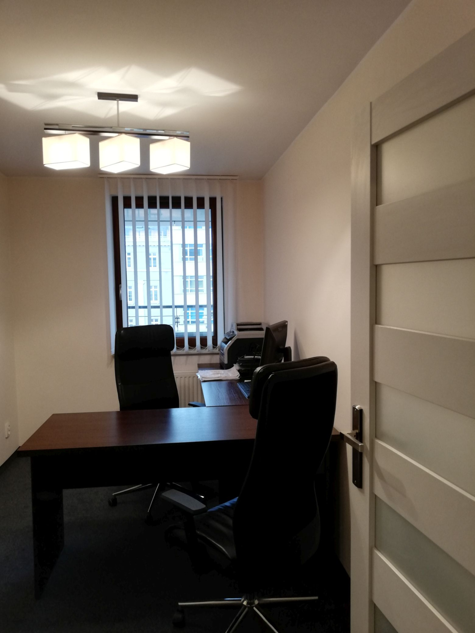 Jasne biuro rachunkowe z biurkiem, krzesłami i oknem z żaluzjami pionowymi. Widok na budynek zza okna. Nowoczesna lampa sufitowa.