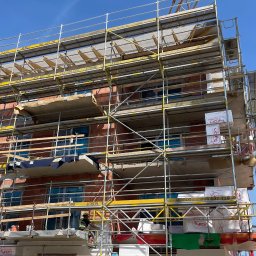 Oleksandr Kalashnyk - Budynek w trakcie budowy, otoczony rusztowaniami, z widocznym dźwigiem w tle. Pracownik w kasku na rusztowaniu. Elementy izolacyjne i materiały budowlane.