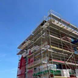 Budynek w trakcie ocieplania, osłonięty rusztowaniami i siatką ochronną na tle błękitnego nieba. Widoczne materiały izolacyjne i elementy konstrukcyjne.