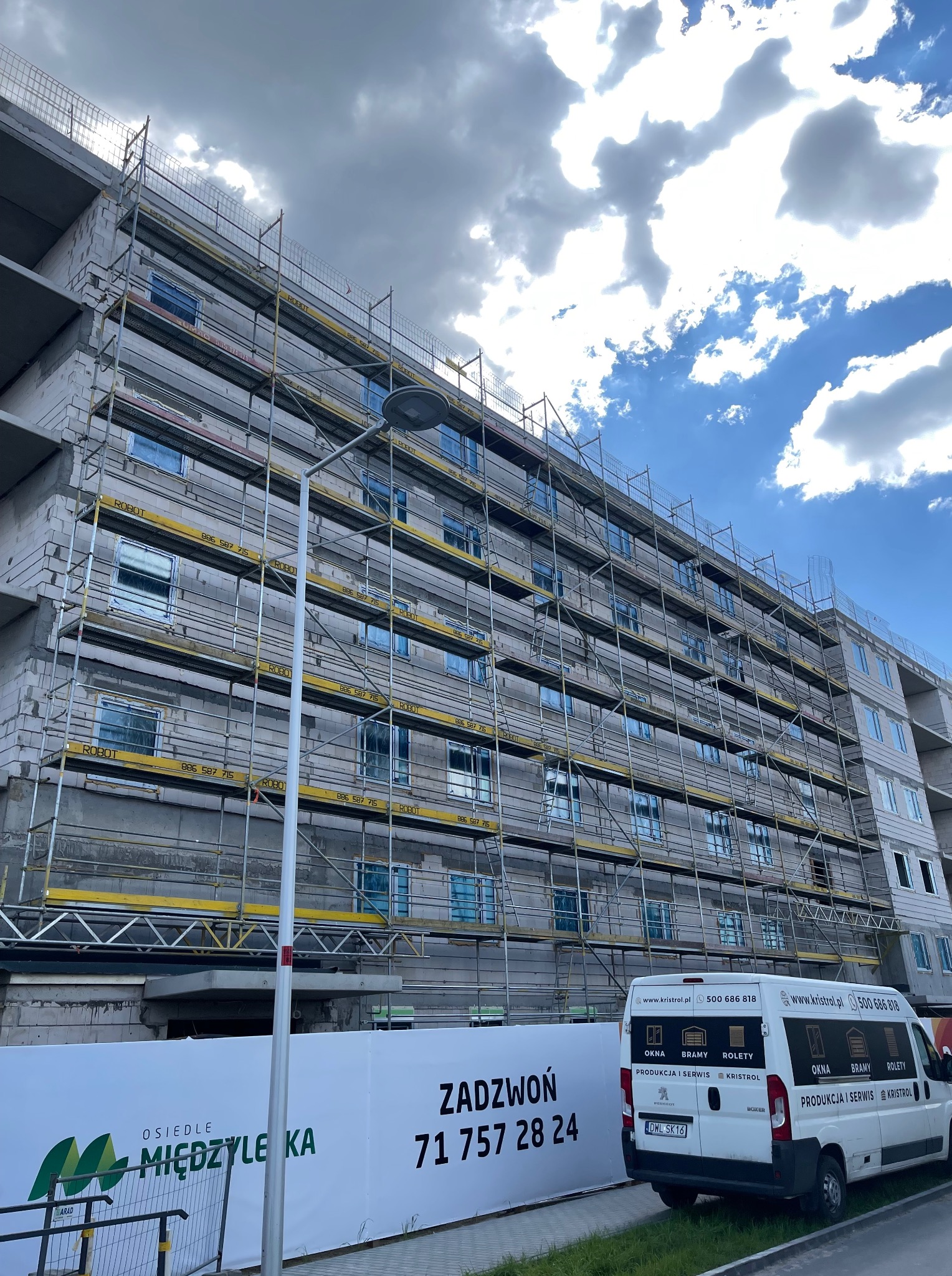 Budynek w trakcie budowy z rusztowaniami, widoczne okna w folii ochronnej, reklama firmy z branży okien i bram na ogrodzeniu i samochodzie dostawczym.