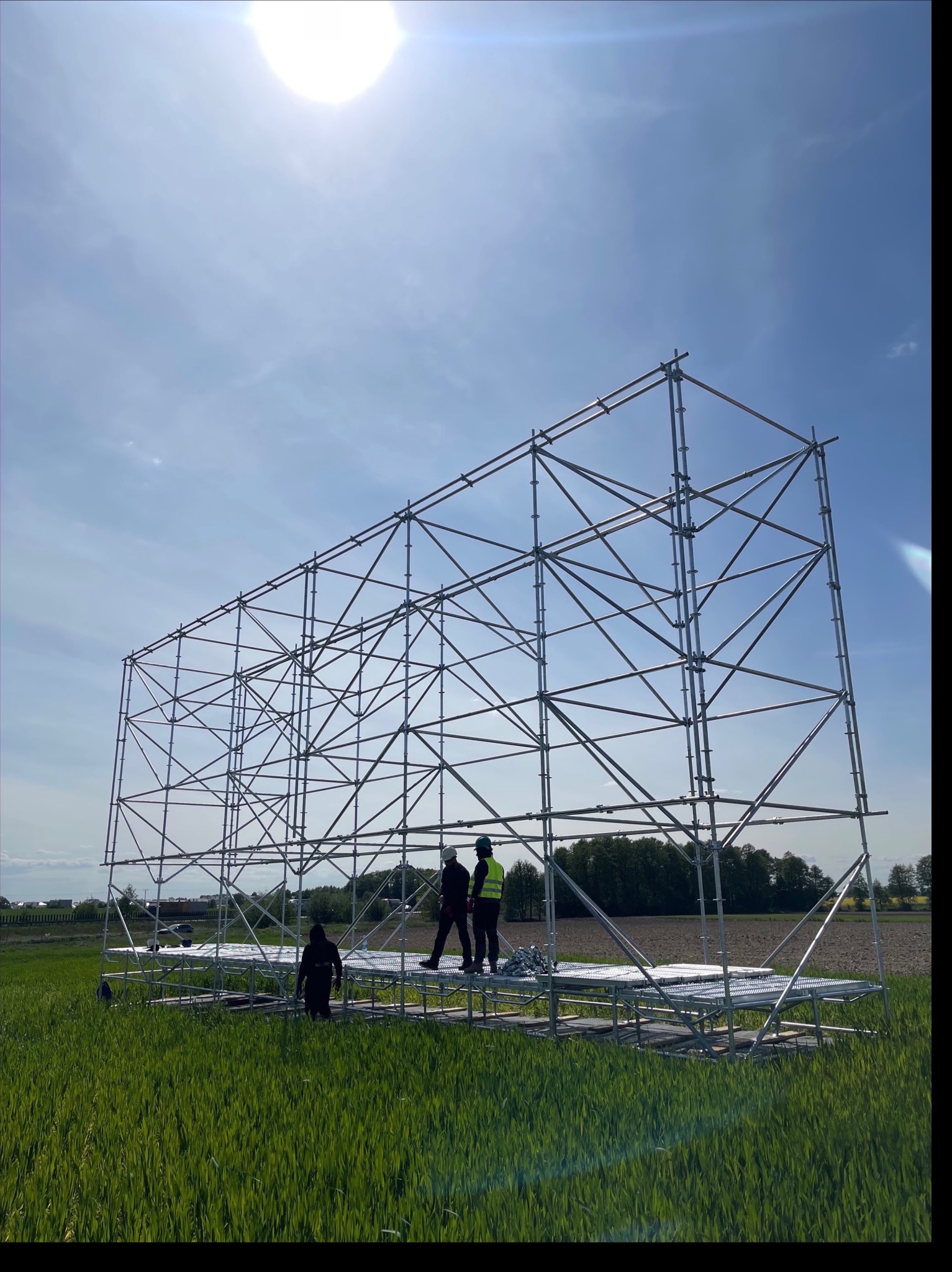 Montaż metalowej konstrukcji rusztowania na zielonej łące w słoneczny dzień. Widoczne osoby w odblaskowych kamizelkach podczas pracy.