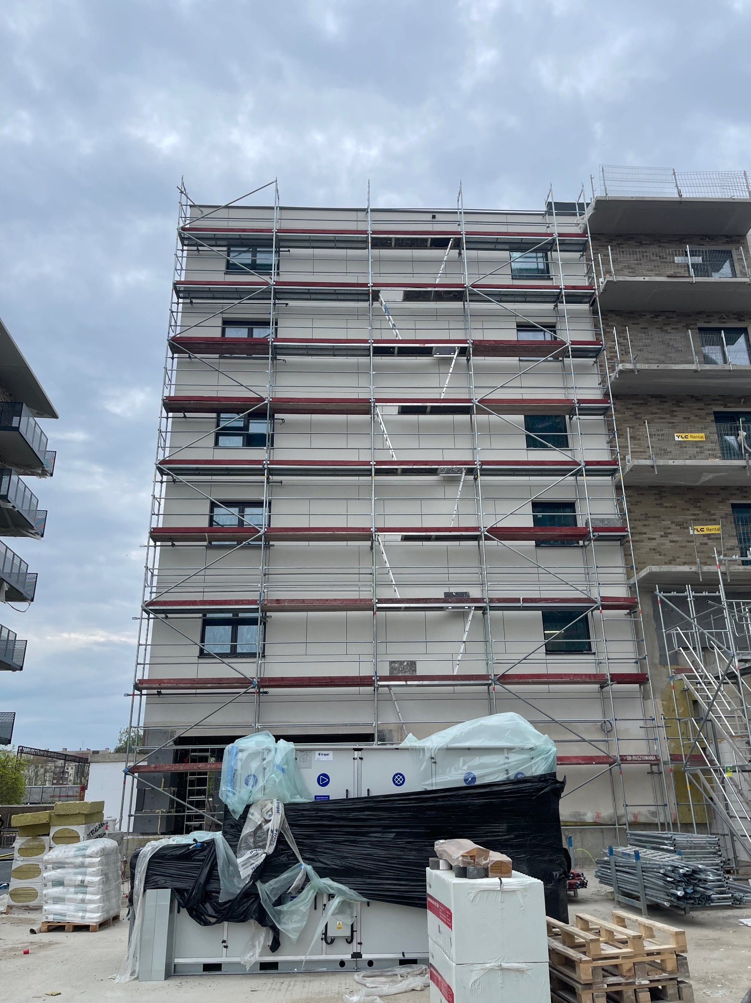 Budynek w trakcie prac elewacyjnych, otoczony rusztowaniami. Widoczne materiały budowlane i sprzęt. Niska perspektywa, pochmurne niebo.