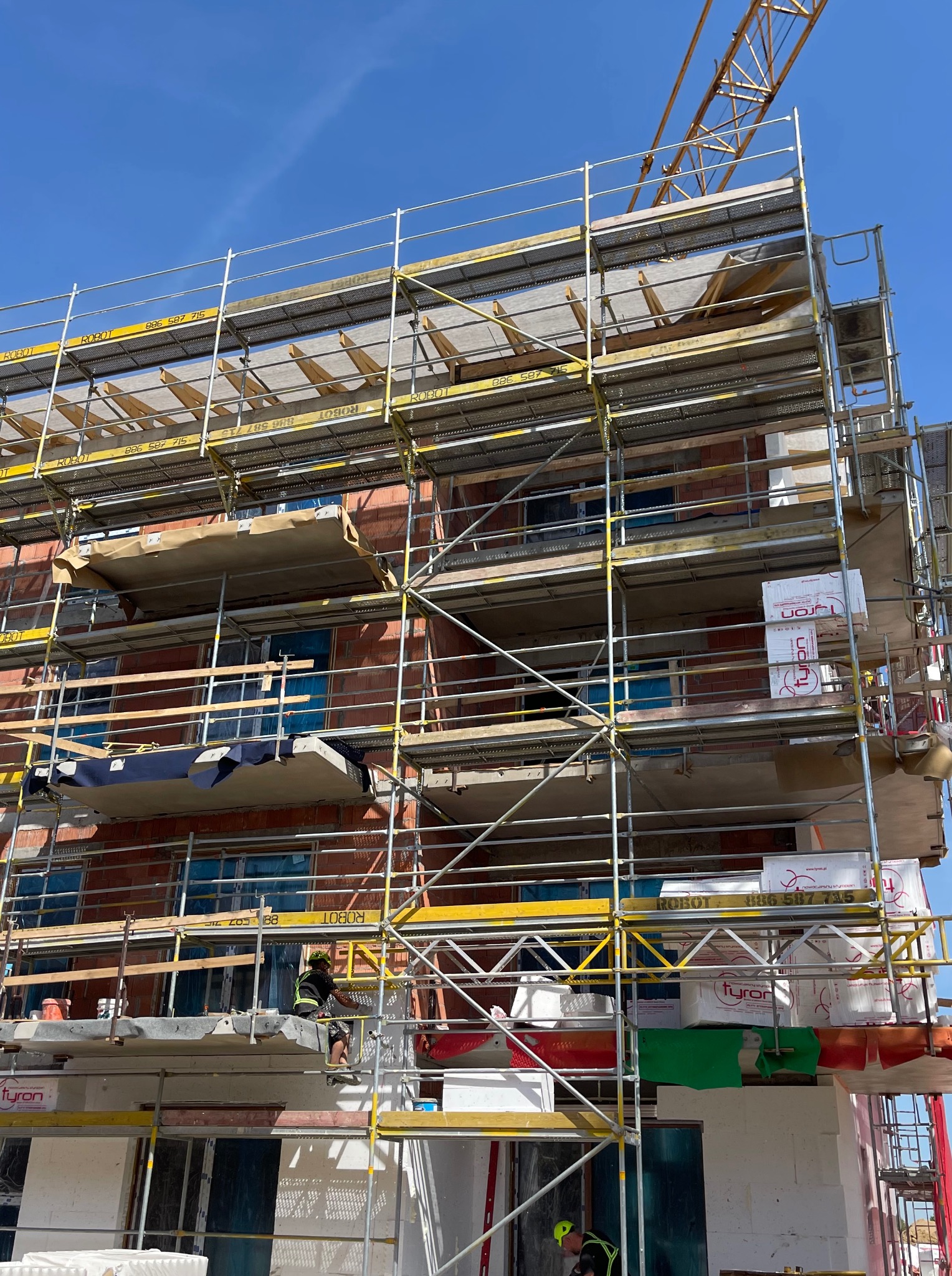 Budynek w trakcie budowy, otoczony rusztowaniami, z widocznym dźwigiem w tle. Pracownik w kasku na rusztowaniu. Elementy izolacyjne i materiały budowlane.