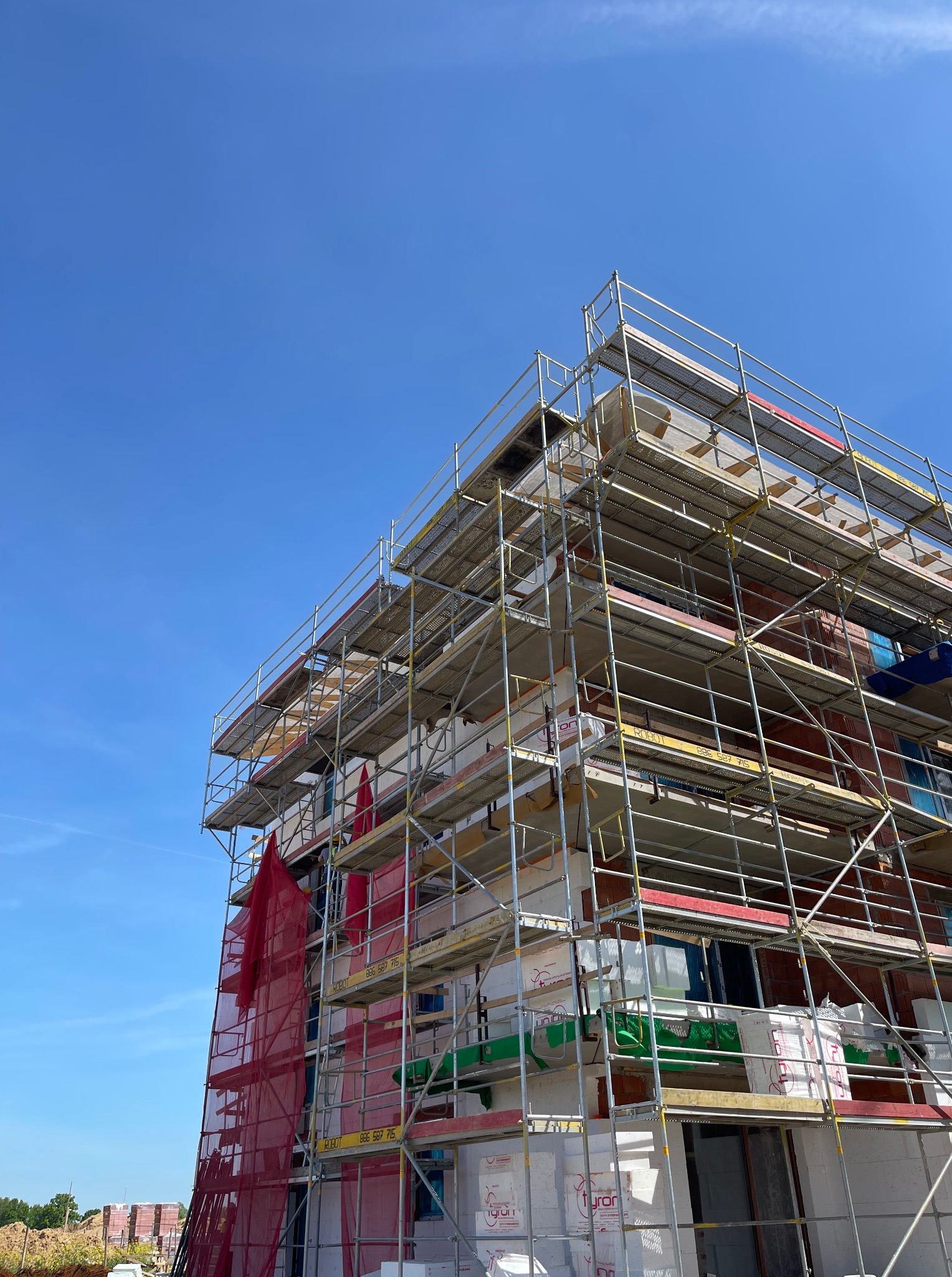 Budynek w trakcie ocieplania, osłonięty rusztowaniami i siatką ochronną na tle błękitnego nieba. Widoczne materiały izolacyjne i elementy konstrukcyjne.