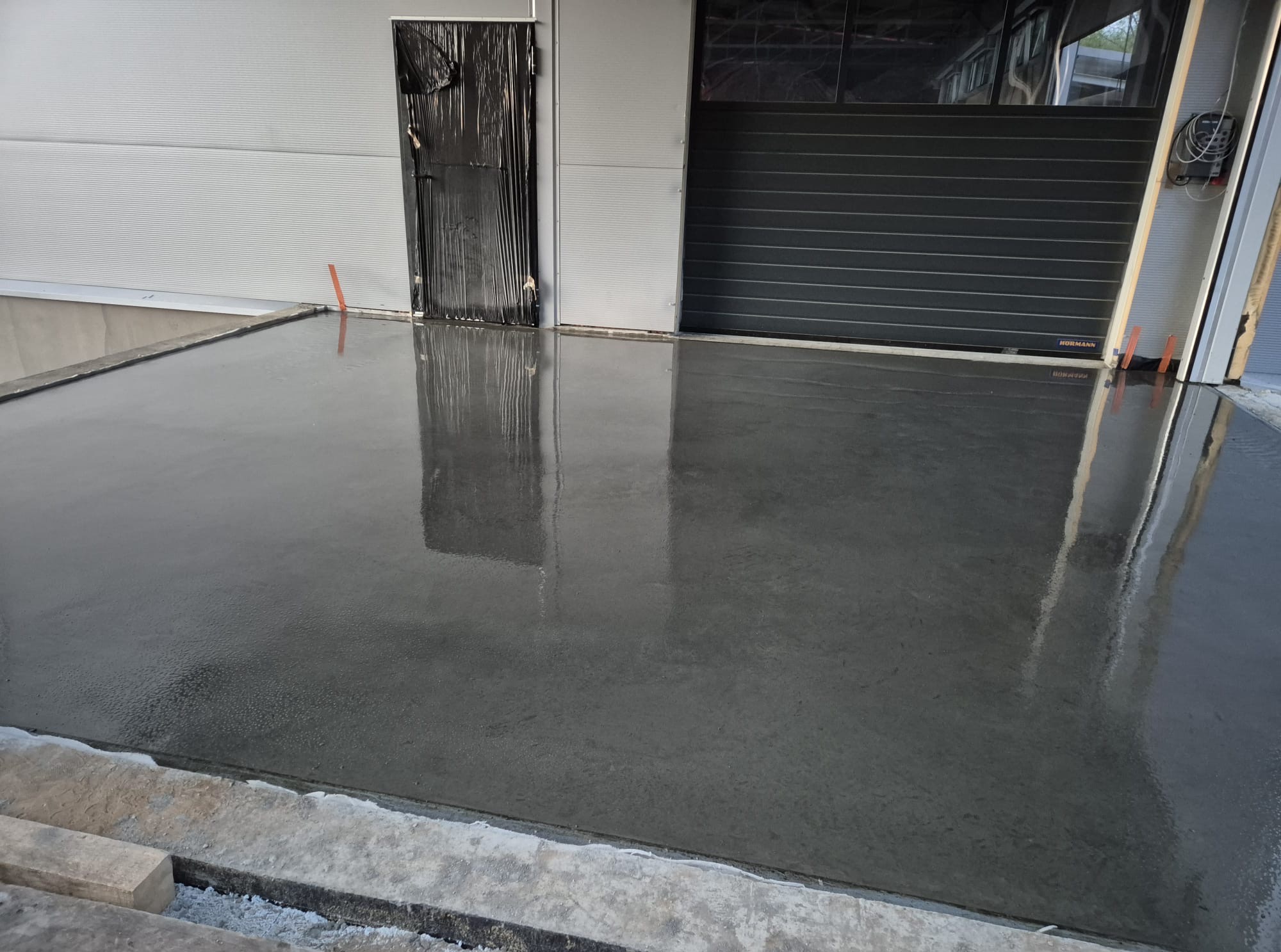 Świeżo wylana, gładka posadzka przemysłowa w hali, z widocznymi refleksami światła. Drzwi i brama garażowa w tle. Widoczny fragment krawężnika.