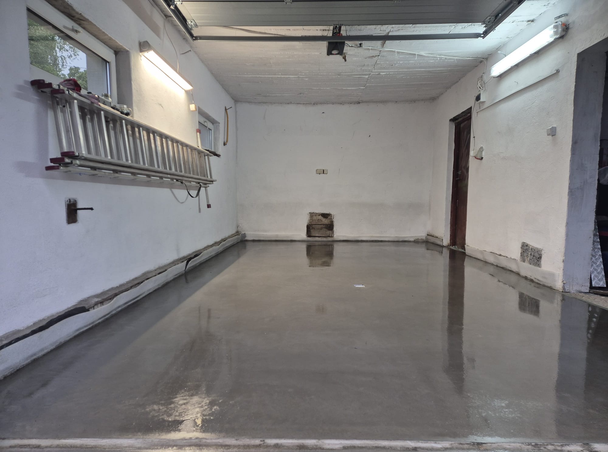 Świeżo wylana, gładka, szara wylewka w garażu z drabiną na ścianie. Widoczne refleksy światła na powierzchni. Surowy, industrialny charakter wnętrza.