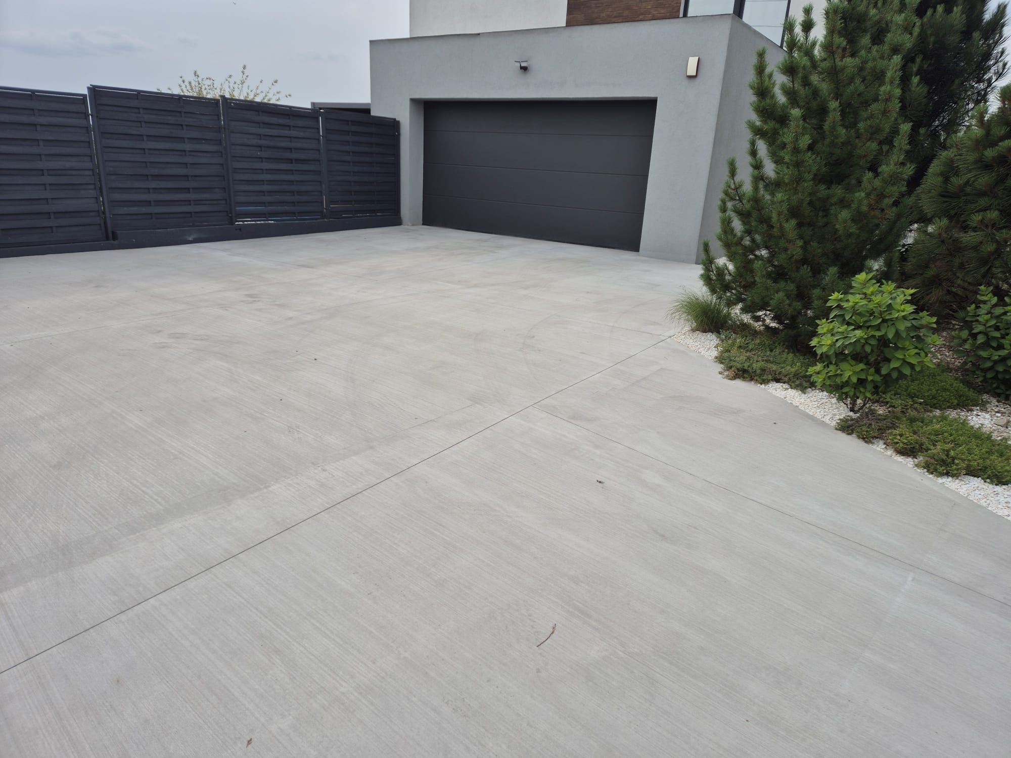 Szara, gładka powierzchnia betonowa podjazdu z widocznymi liniami dylatacyjnymi, prowadząca do garażu nowoczesnego domu z ciemną bramą i ogrodzeniem.