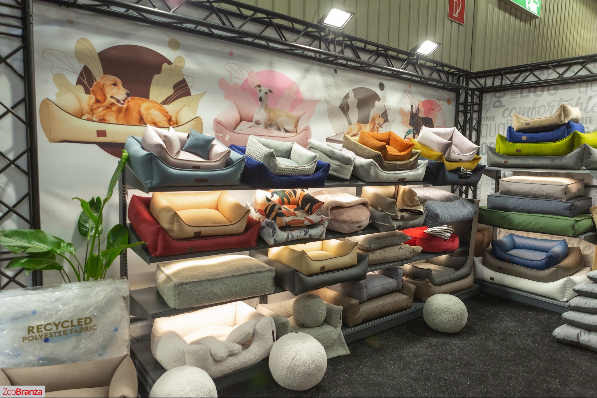 Ekspozycja różnorodnych, kolorowych legowisk dla psów na metalowych półkach. W tle grafiki psów w legowiskach i logo firmy ZooBranza. Widoczny napis: Recycled Polyester Fabric.