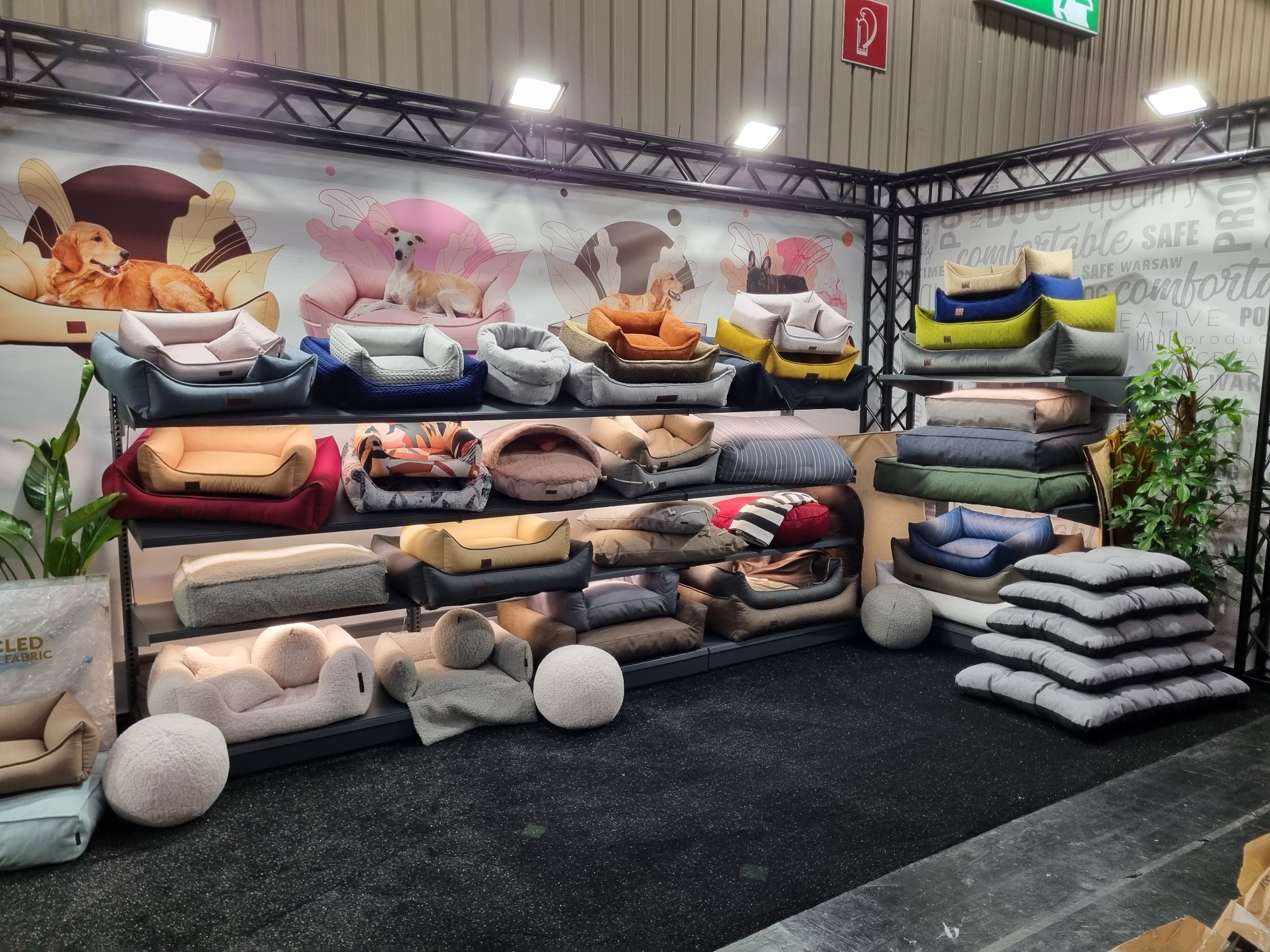 Ekspozycja różnorodnych legowisk dla psów na półkach, z grafikami psów w tle, prezentowana na targach. Widoczne różne style, kolory i materiały.