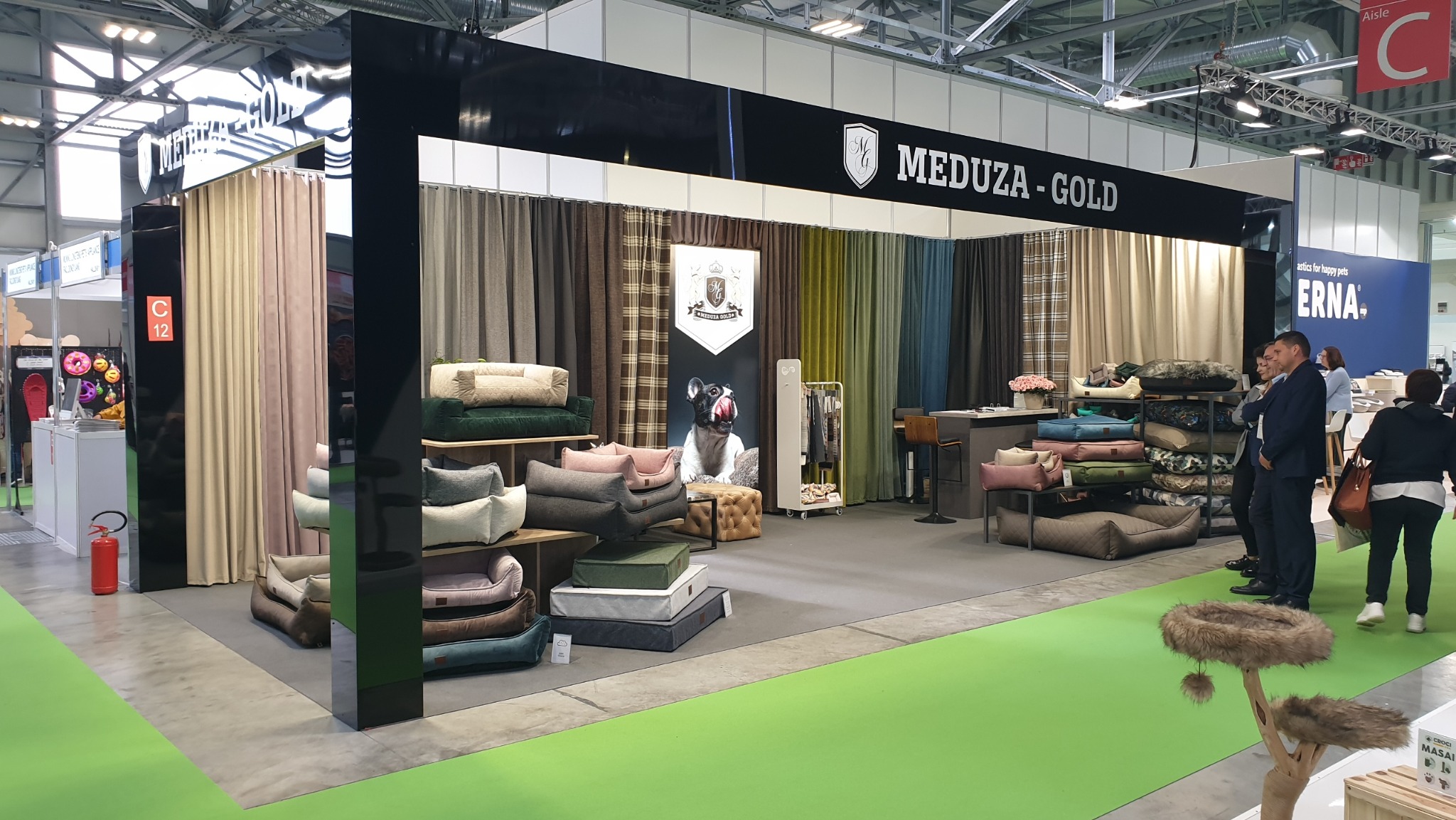 Ekspozycja stoiska Meduza-Gold na targach: różnorodne legowiska dla zwierząt, zasłony w tle, osoby w garniturach, zielona podłoga. Prezentacja produktów.