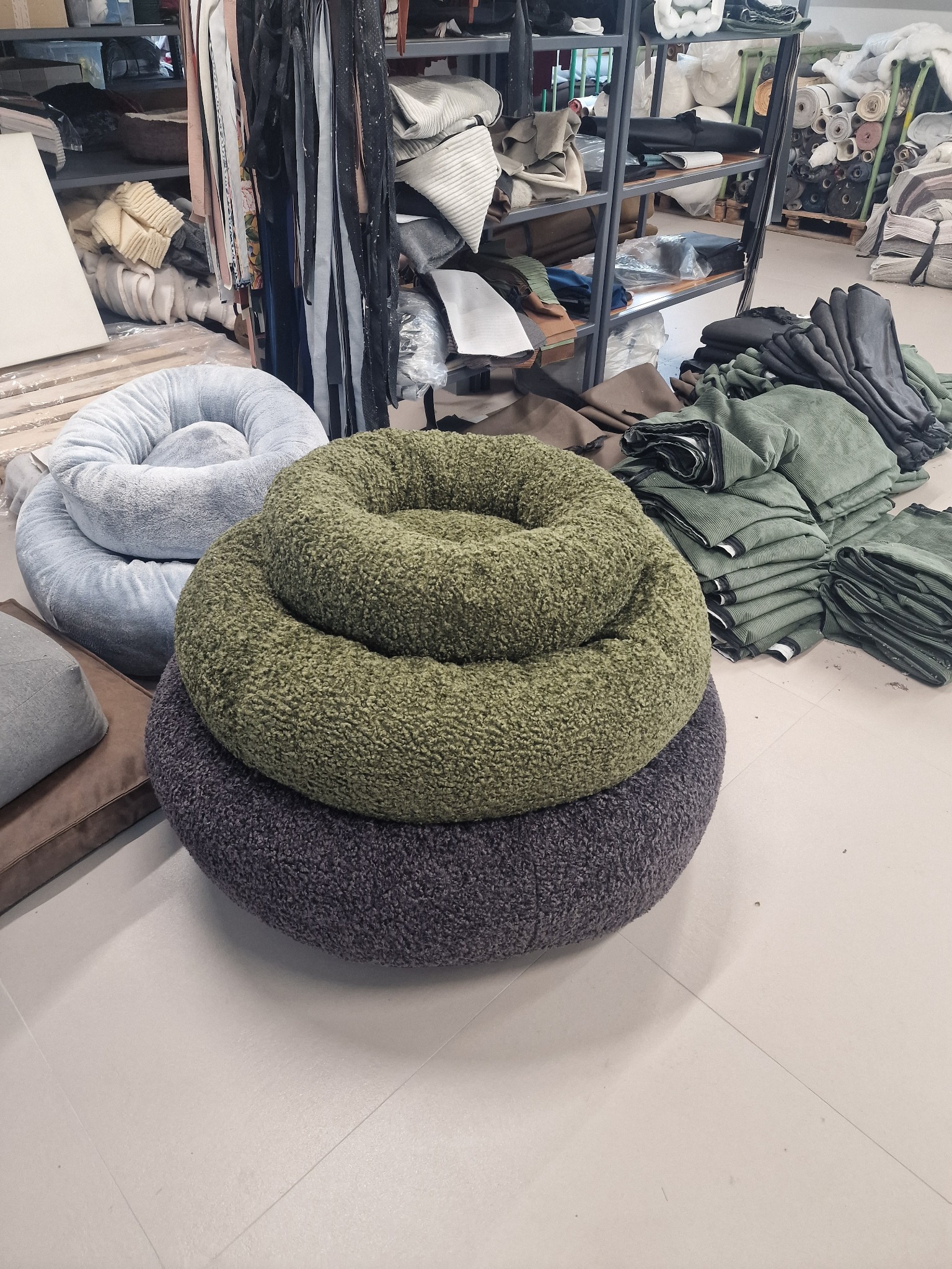 Trzy okrągłe, teksturowane legowiska dla zwierząt w odcieniach szarości, zieleni i fioletu, ułożone jedno na drugim w szwalni z tkaninami na półkach w tle.