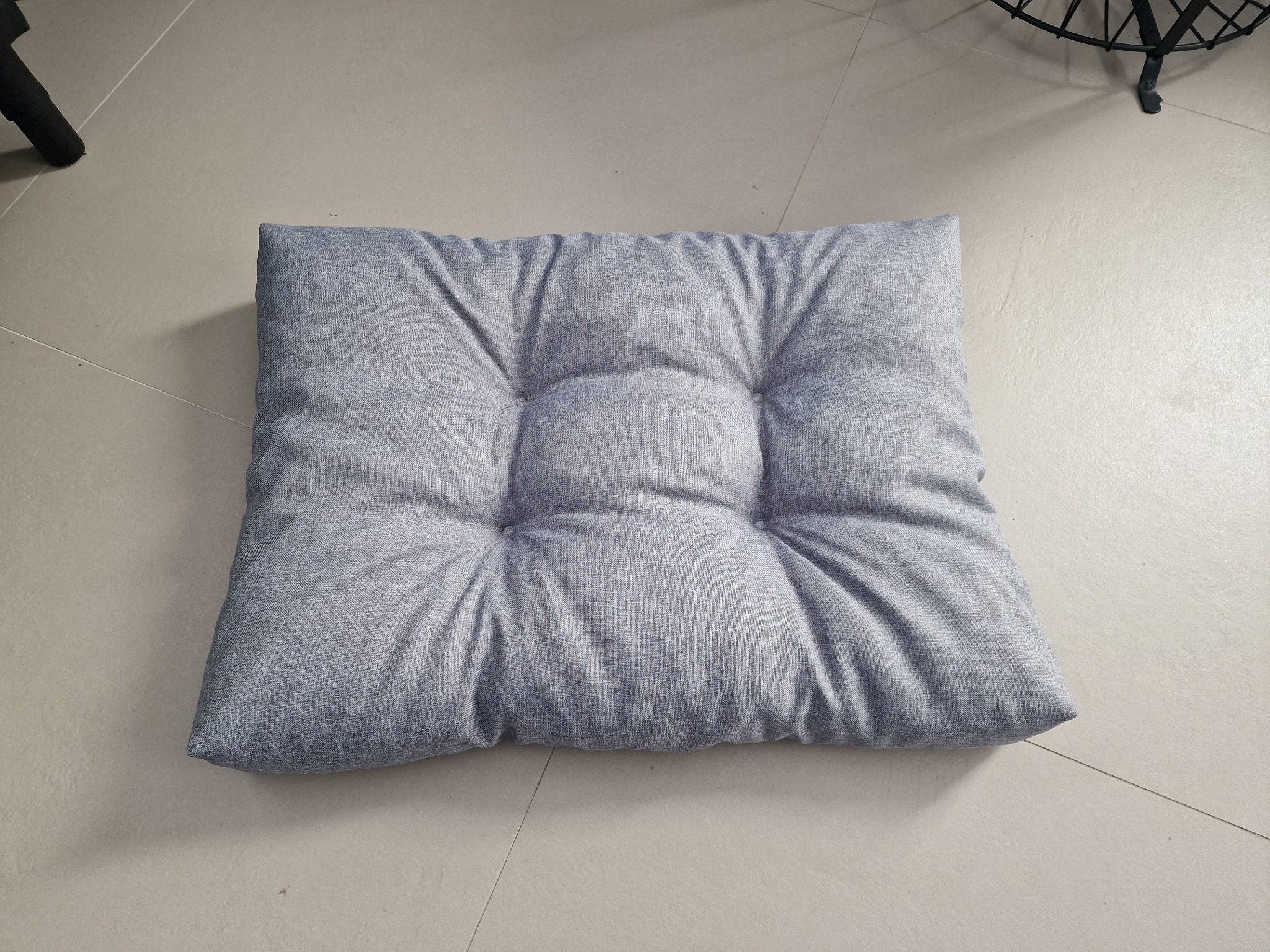 Szara, kwadratowa poduszka na krzesło z sześcioma przeszyciami na jasnej, kafelkowanej podłodze. W tle fragment czarnego krzesła.