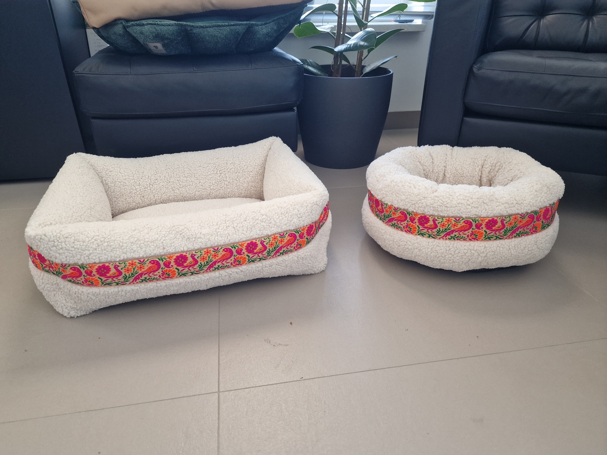 Dwa białe, pluszowe legowiska dla zwierząt z kolorową taśmą z motywem kwiatowym i ptasim, na tle szarej podłogi i ciemnych mebli.